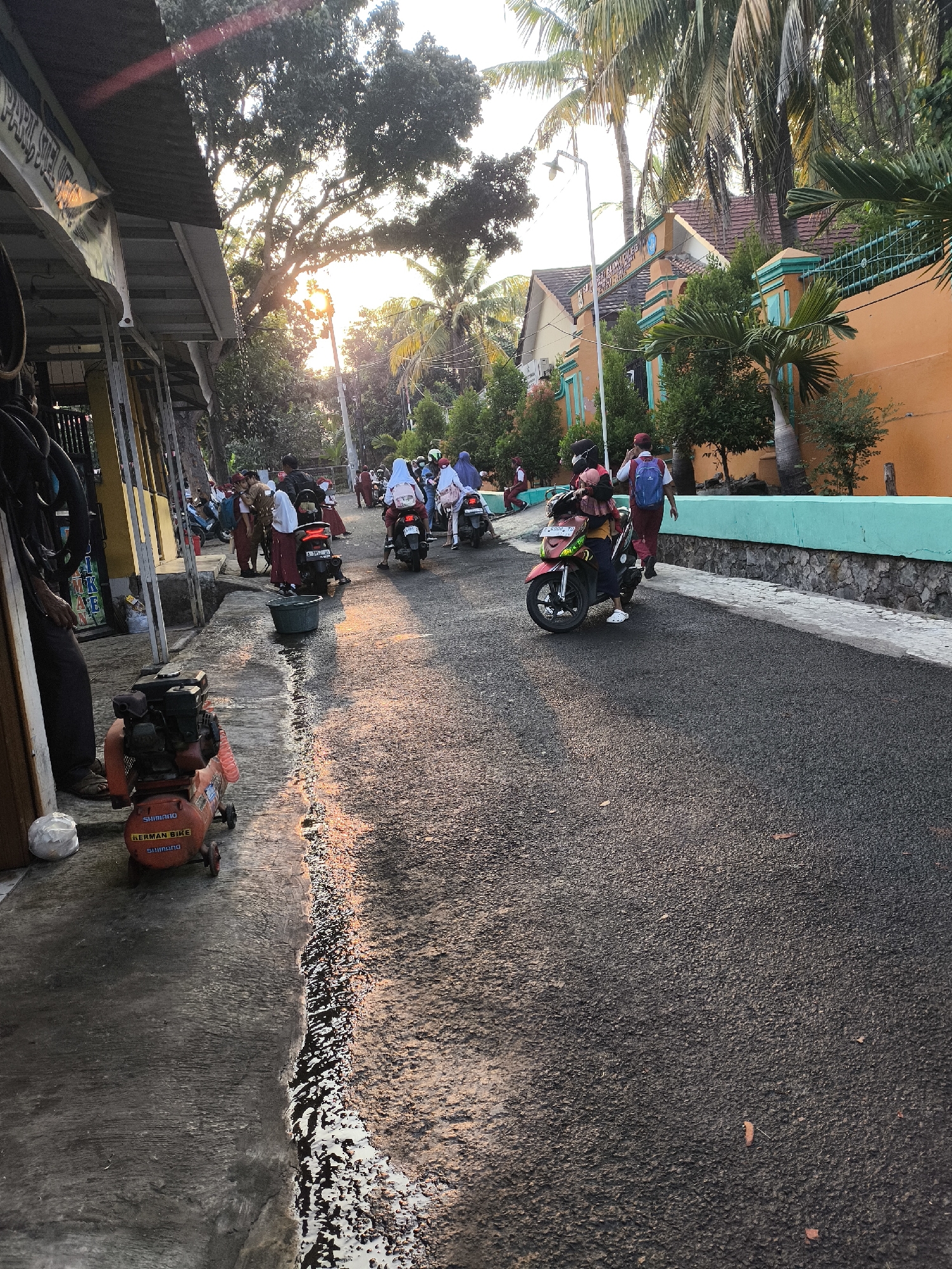 The image depicts a street scene in Cilegon, Indonesia, likely in the late afternoon given the warm, angled sunlight illuminating the right side of the asphalt road and the shadows cast to the left. Several individuals, predominantly young students wearing school uniforms consisting of red and white attire, are present. Some are on foot, while others are riding motorcycles. One person on a motorcycle, a student with a red backpack, is prominently featured in the foreground. The street is lined with buildings on the left and right. On the left, a covered walkway or shop front with signage is visible, with a red air compressor situated on the adjacent concrete sidewalk. The right side of the street features an orange building with turquoise accents and a stone wall with a turquoise railing. Palm trees and other foliage are interspersed throughout the scene, suggesting a tropical or subtropical climate. The road itself appears to be paved with dark asphalt and shows signs of recent wear or construction, with a distinct line separating the darker, wet-looking asphalt from the drier, lighter concrete on the left. The overall atmosphere suggests a busy but ordinary moment in a residential or school area of Cilegon.