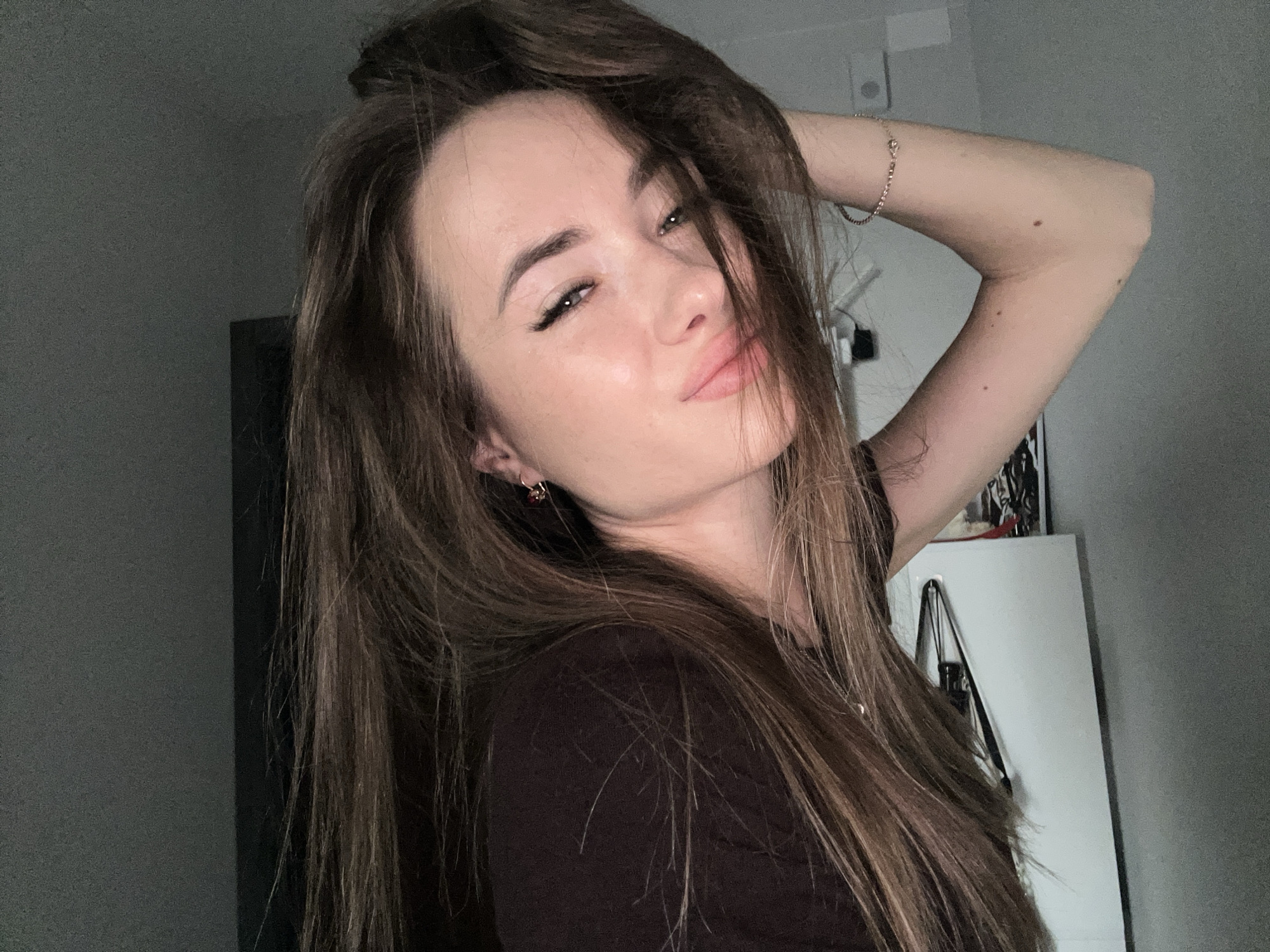 A woman is captured in a selfie, her face angled slightly towards the camera with her right arm raised and her hand touching her hair. She has long, brown hair that partially obscures her face and shoulders. Her eyes are partially closed, and her lips are slightly parted, giving a pouty expression. She is wearing a dark brown, short-sleeved shirt and a thin bracelet on her right wrist. The background is a plain, grey wall, with a white cabinet visible to the right. The lighting is dim, creating shadows on her face.