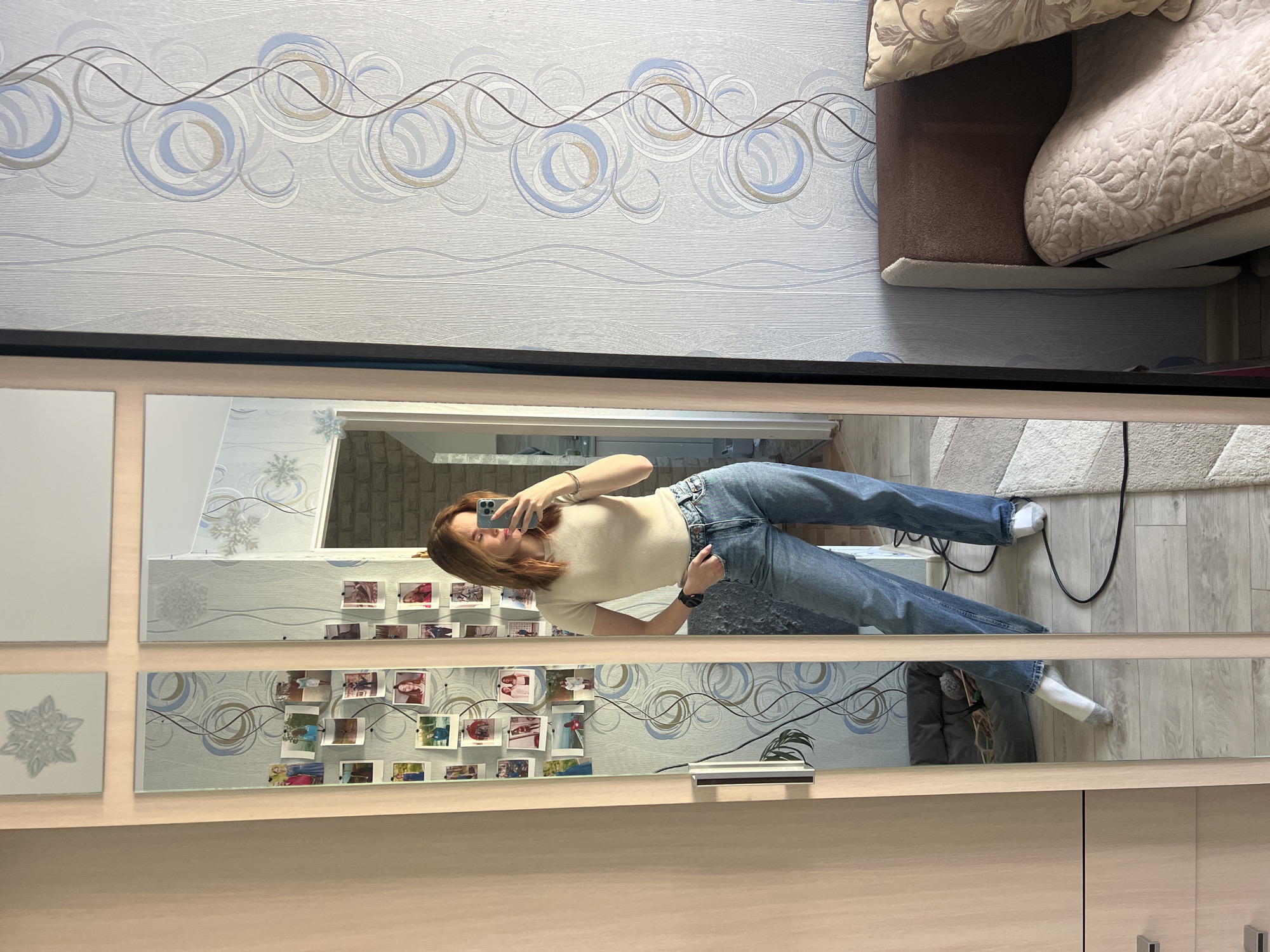 A woman is taking a picture of herself in a mirror. The mirror is mounted on a piece of furniture and the woman is standing in front of it. The woman is wearing a light-colored shirt and blue jeans. She is holding a phone in her right hand. The mirror is reflecting the room behind her. The room has patterned wallpaper and a couch in the upper right corner. There are photographs attached to the mirror on the lower left. The location is Chaikovsky, Russia.
