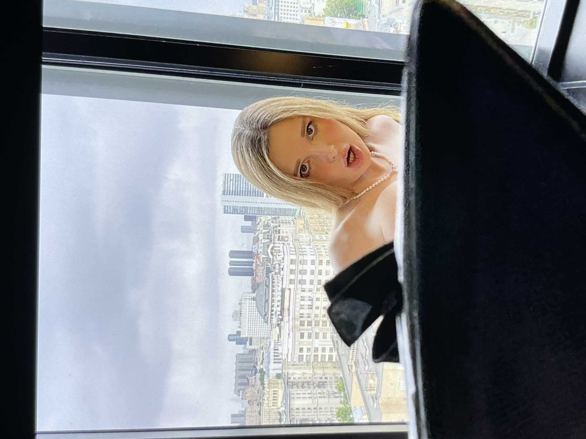 A young woman with blonde hair and makeup is leaning into a window, looking at the camera with her mouth open. She is wearing a pearl necklace. Behind her, a city skyline is visible under a cloudy sky. In the foreground, a black object, possibly furniture, is partially obscuring the view.