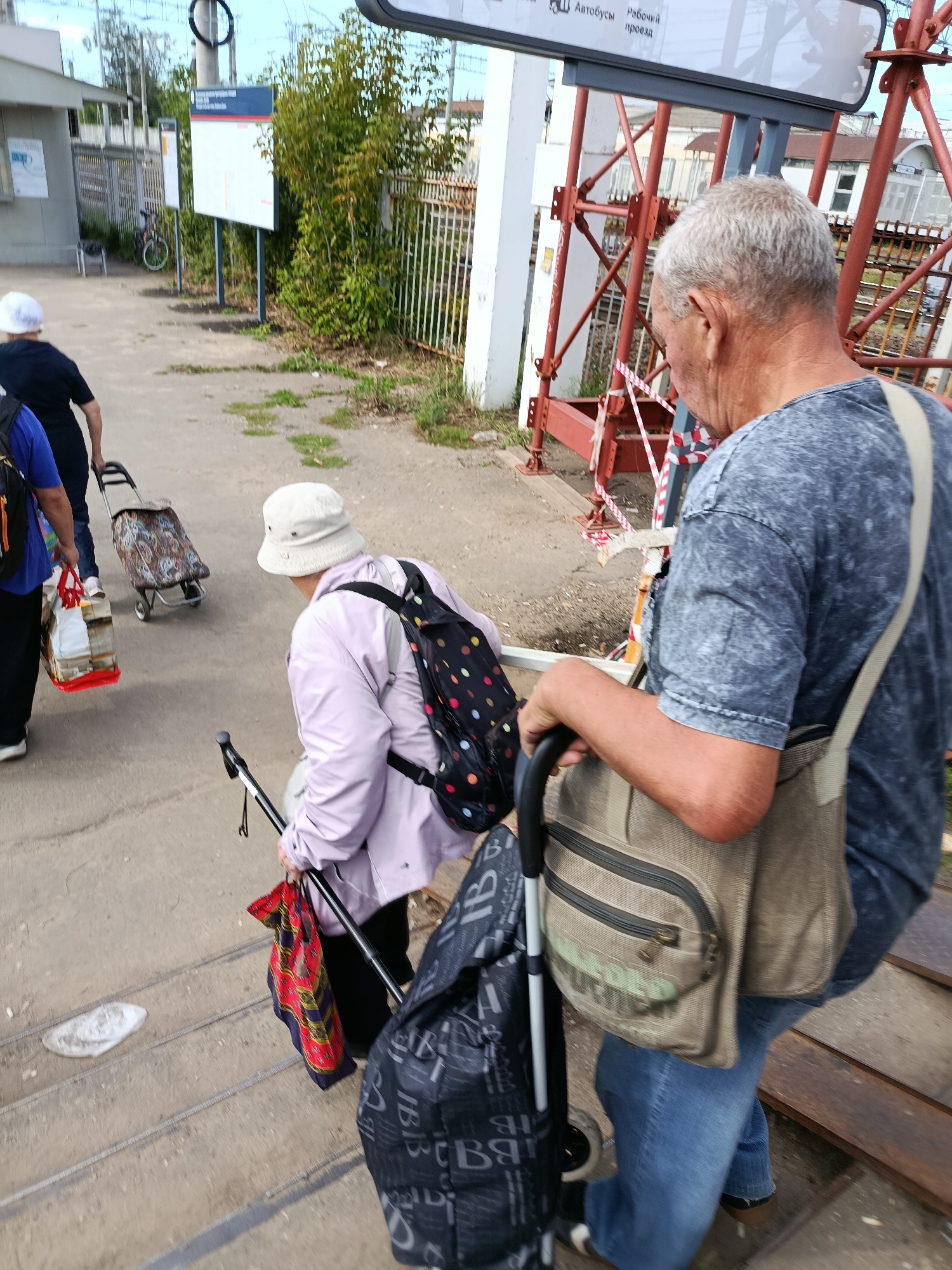 At a train station in Orekhovo-Zuyevo, Russia, an elderly man with grey hair wearing a grey t-shirt and blue jeans walks up a ramp, carrying a tan messenger bag. He is followed by an elderly woman in a light pink jacket and a light-colored hat, who is also walking up the ramp and using a walking stick. She has a polka-dotted backpack and is holding a colorful scarf. Behind them, another person in a blue t-shirt is pulling a shopping cart with bags. The environment includes a train platform, railway tracks, a fence, and signage.