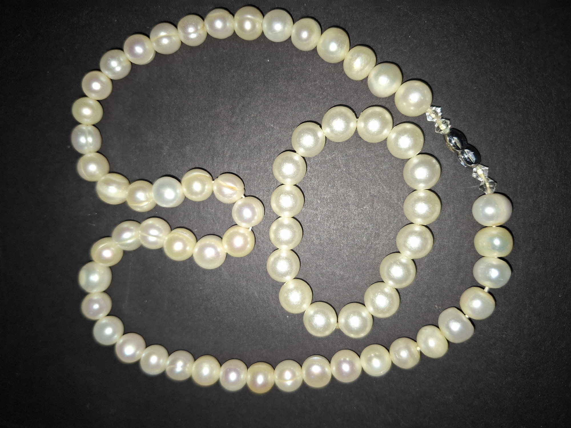 A pearl necklace and bracelet are laid out on a dark surface. The necklace is a strand of irregularly shaped, off-white pearls, arranged in a loose figure-eight shape. The bracelet, also made of similar pearls, is coiled and lies beneath the necklace. Both pieces feature a clasp composed of clear, faceted beads and dark, iridescent beads. The lighting creates subtle shadows and highlights on the pearls, emphasizing their luster. The background is a uniform dark color, likely a table or cloth, providing a stark contrast to the jewelry. The scene is entirely devoid of any indication of the city of Almaty, Kazakhstan or any urban environment.