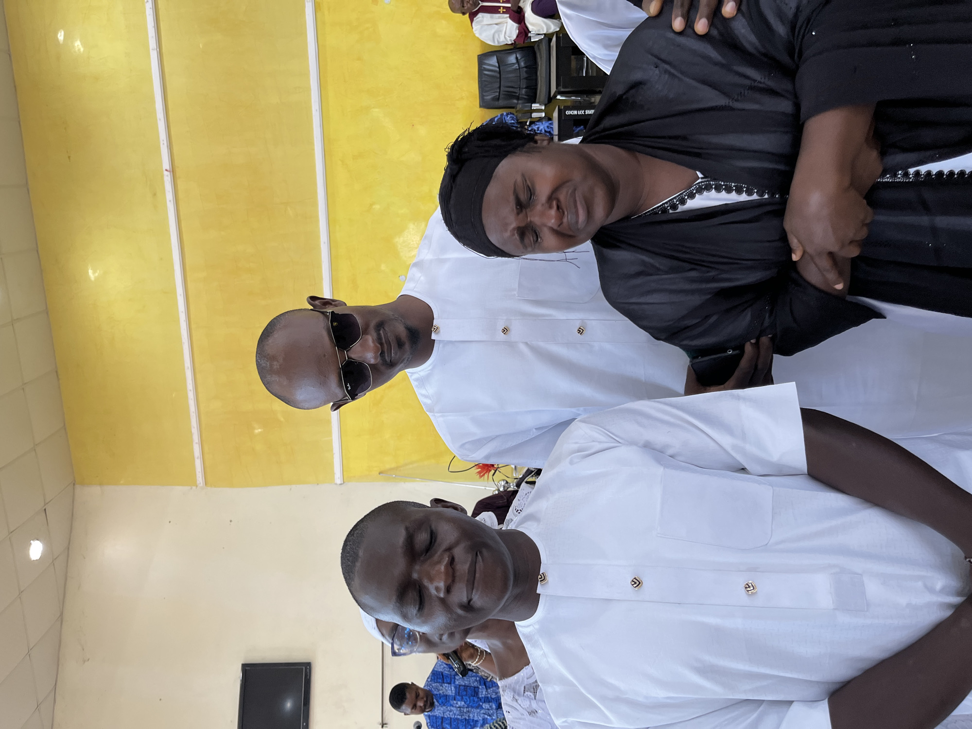 The image shows a group of people, possibly at an event or gathering. A woman with a black head wrap and a black and white dress, stands near a man in sunglasses and a white shirt. Another man in a white shirt stands in front. The background includes a yellow wall and chairs with other people in the distance. The location is likely indoors, but there is no specific evidence to confirm the city of Jos, Nigeria.