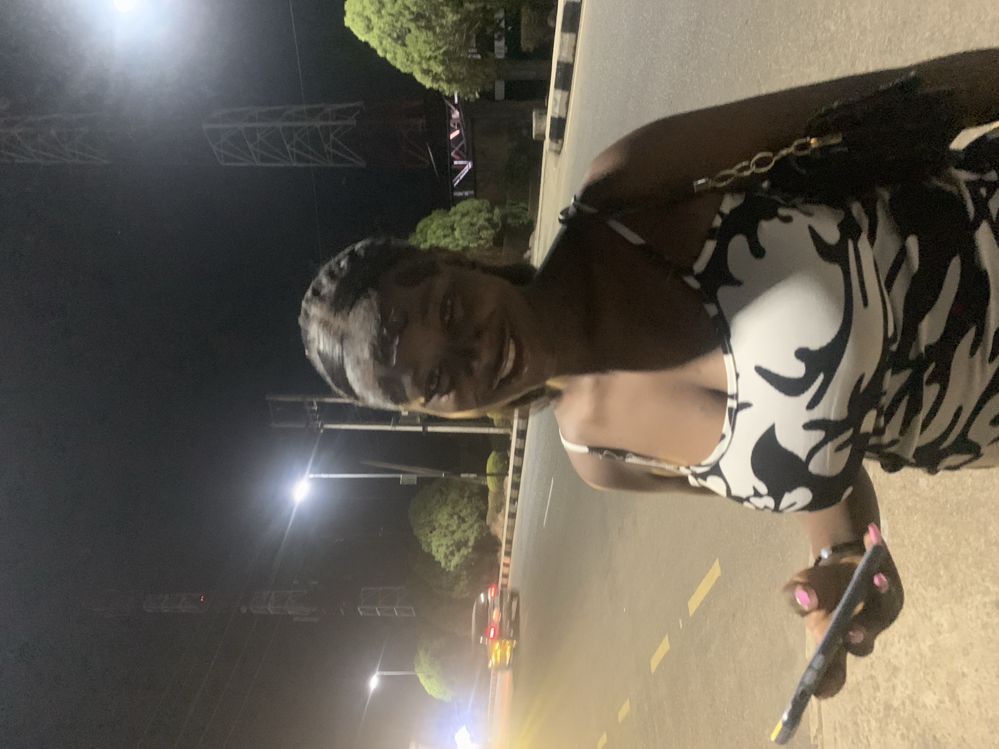 A medium shot of a dark-skinned woman holding a phone while standing on a street at night. The woman is wearing a black-and-white patterned dress with thin straps, and a black purse hangs over her shoulder. Her fingernails are painted pink. She is smiling slightly and looking at the camera. The background includes streetlights, trees, a median with black-and-white striped barriers, and a road with yellow lane markings. In the distance, there is a car with its headlights on, and tall metal structures are visible above the trees. The location is identified as Jos, Nigeria, due to the specific architecture and street design elements found in the background.