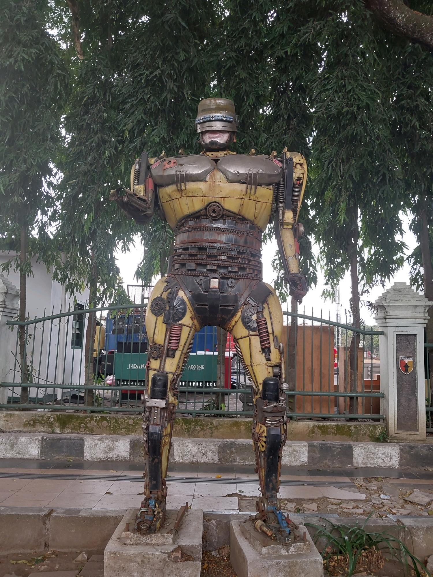 A full-body, life-sized sculpture of Robocop, constructed from salvaged metal and mechanical parts, stands outdoors on a paved sidewalk. The sculpture is weathered and appears to be made from a variety of scrap materials, including car parts, gears, chains, and springs. The sculpture is positioned in front of a metal fence and several trees with lush green foliage. Behind the fence, two trucks are partially visible. The sidewalk has a decorative pattern of black and white concrete blocks. A stone pillar with a crest is visible to the right of the sculpture. The background suggests an urban environment, possibly a park or public space in the city of Malang, Indonesia, given the presence of a crest with the text "SAKA BHAYANGKARA". The sculpture is a static display, with no people interacting with it. The lighting suggests it is daytime, with diffused light filtering through the trees.