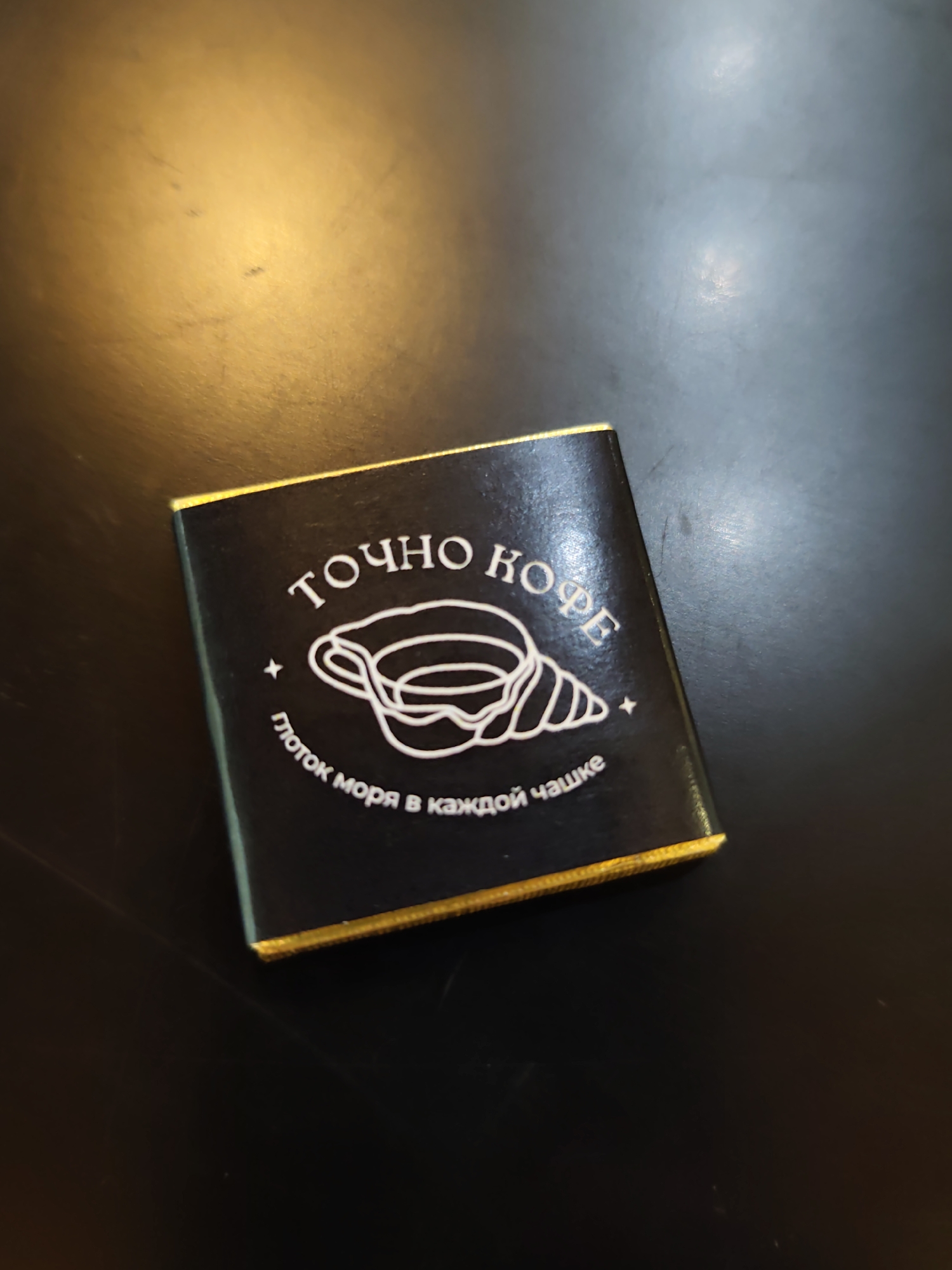 This is a top-down, eye-level shot of a dark, glossy surface, possibly a table. The surface has some minor scuffs and is illuminated by a warm light source from the upper left, creating a gradient of light and shadow.
Dominating the center of the frame is a small, black, rectangular object with a gold foil border. This object is likely a chocolate wrapper. The wrapper features white text and an illustration. The text in a curved arc at the top reads "ТОЧНО КОФЕ," which translates to "EXACTLY COFFEE." Below this text is a stylized white illustration of a conch shell or a similar spiral form within a cup shape. Underneath the illustration, another line of white text curves around: "глоток моря в каждой чашке," translating to "a sip of the sea in every cup."
The composition is minimalist, with the subject centered and the dark background providing a stark contrast. There are no people or animals present in the image, and no specific landmarks or indications of the city of Sochi, Russia, are visible. The environment appears to be indoors, given the controlled lighting and the nature of the surface.