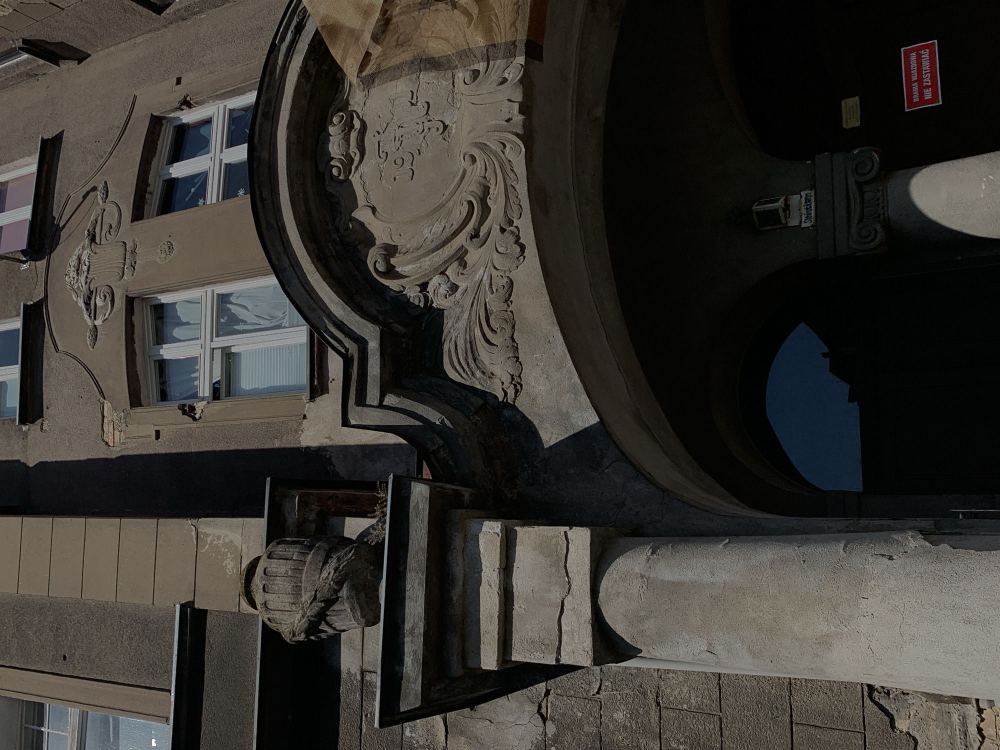 The image captures the facade of a historical building in Poznan, Poland. A stone archway dominates the lower portion of the frame, leading into a darkened interior. Above the arch, intricate stone carvings adorn the building's exterior, including a decorative crest and what appears to be a date carved into the stone. To the left of the arch, a cylindrical stone pillar supports an overhang, upon which rests a decorative stone urn with rope-like carvings. The building's facade is constructed of textured plaster and stone, showing signs of age and wear, with visible cracks and crumbling sections. Sunlight casts strong shadows across the building, highlighting the architectural details. A red sign with white text is visible within the archway, bearing the Polish inscription "BRAMA WJAZDOWA NIE ZASTAWIAĆ," which translates to "Entrance Gate Do Not Block." The overall impression is of an old, possibly monumental, structure in an urban setting.