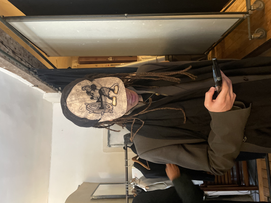 A person wearing a Mickey Mouse hat and a layered jacket holds a phone. They have dreadlocks. Behind them are clothes hanging on a rack and a large whiteboard with a metal frame. The background also includes a wall and some wooden flooring. The image seems to be taken indoors.
