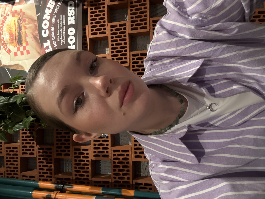 A young woman with her hair pulled back poses in a lavender and white striped shirt. She wears a green beaded necklace and a silver crescent moon pendant. Behind her is a brick wall and a menu board advertising food items, including a "double cheese burger" and "basil lemonade", priced at 400 RSD. There are some potted plants visible in the corner.