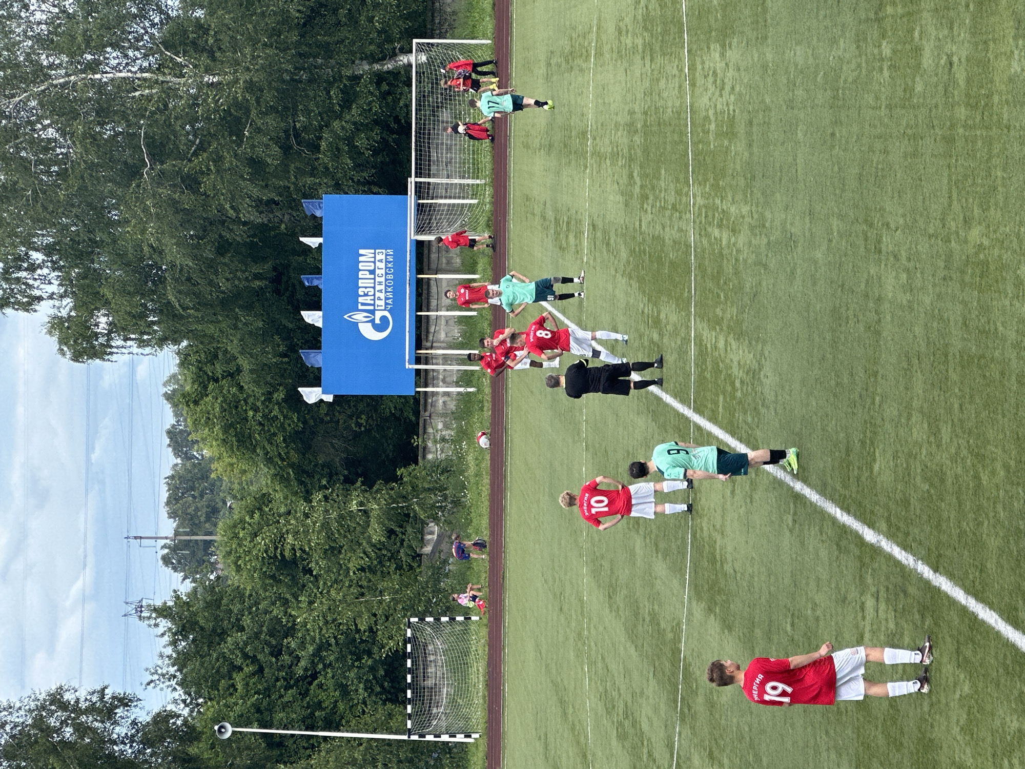 The image depicts a soccer match on a green artificial turf field. In the background, a blue banner with the Gazprom logo and text in Russian, "ГАЗПРОМ ТРАНСГАЗ ЧАЙКОВСКИЙ," is visible, indicating the location as Chaikovsky, Russia. Several players are scattered across the field, some in red jerseys with numbers and white shorts, others in green jerseys and dark shorts, and one in a black referee uniform. Two players in red jerseys, one with the number 10 on his back and another with the number 19, are prominently featured. A player in a green jersey with the number 9 is also visible. In the far background, a soccer goal with a net is set up. The sky is overcast with some clouds, and a line of trees forms a backdrop to the scene.