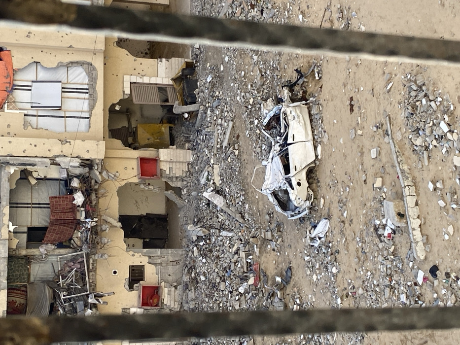 This image depicts a scene of extensive urban devastation in Khan Yunis, Palestinian Territory, viewed from an elevated position, partially obstructed by two blurry vertical bars in the foreground. The primary subject is a severely damaged, light-colored, multi-story building occupying the background. Its facade is heavily pockmarked with numerous small impact craters, indicative of bullet or shrapnel damage. Large sections of the ground floor walls have collapsed, revealing dark, debris-filled interiors; the left opening shows two red cube-like structures on the remaining wall, while the right opening exposes a yellow object and a louvered metal door. The upper floors of the building also exhibit significant structural damage, with collapsed sections exposing tangled rebar, scattered household items such as patterned textiles, and what appears to be a metal frame on the left. A window on the upper right is covered with white material secured by dark straps, and an orange textile hangs nearby.
The ground area between the viewer and the building is covered in a dense mixture of sand, dirt, and significant amounts of concrete and masonry rubble. In the mid-ground, a heavily damaged white four-door sedan rests on the sandy terrain. The vehicle is crumpled and appears largely flattened, with its roof collapsed, windows missing, and body panels severely distorted, indicating a catastrophic impact. Smaller fragments of debris are scattered around the car. To the lower left, a broken concrete barrier or low wall is visible amidst the rubble and sand. No people are present in the scene.