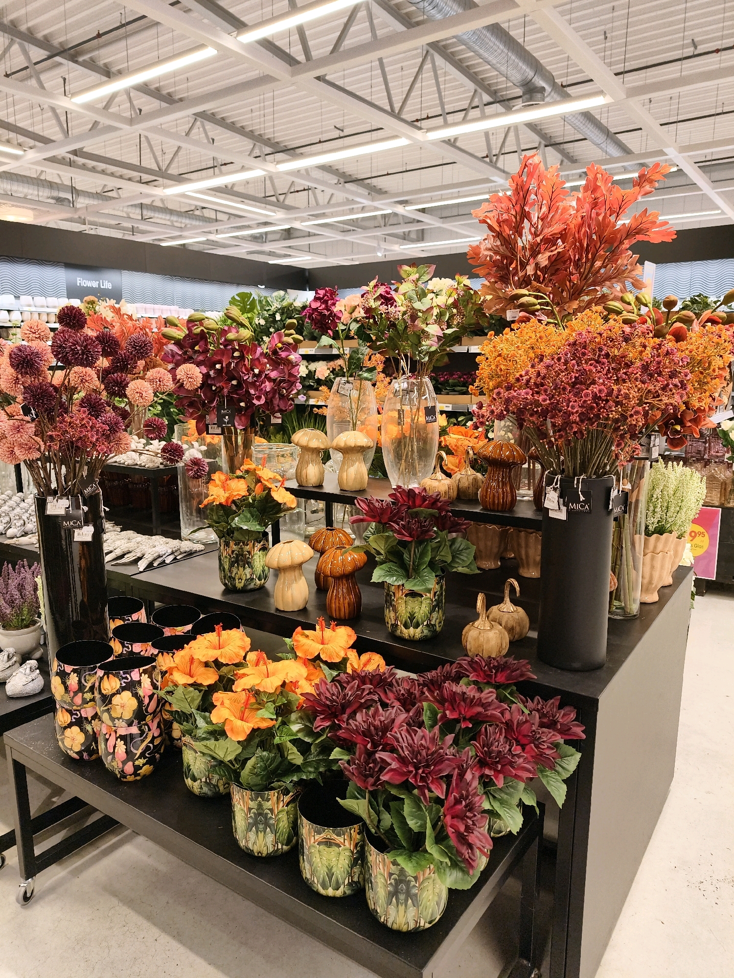 The image shows an indoor display of artificial flowers and decorative items, likely in a retail store. The display features a variety of floral arrangements in different vases, as well as decorative pumpkins and mushroom-shaped objects. The lighting suggests an overhead retail lighting system. The backdrop shows a gray wall with the words "Flower Life" visible. The overall scene is organized and presented for sale. This setup is consistent with what might be found in a store like IKEA in Linköping, Sweden, which is known for its home furnishings and decorative items. There are no people visible in the image.