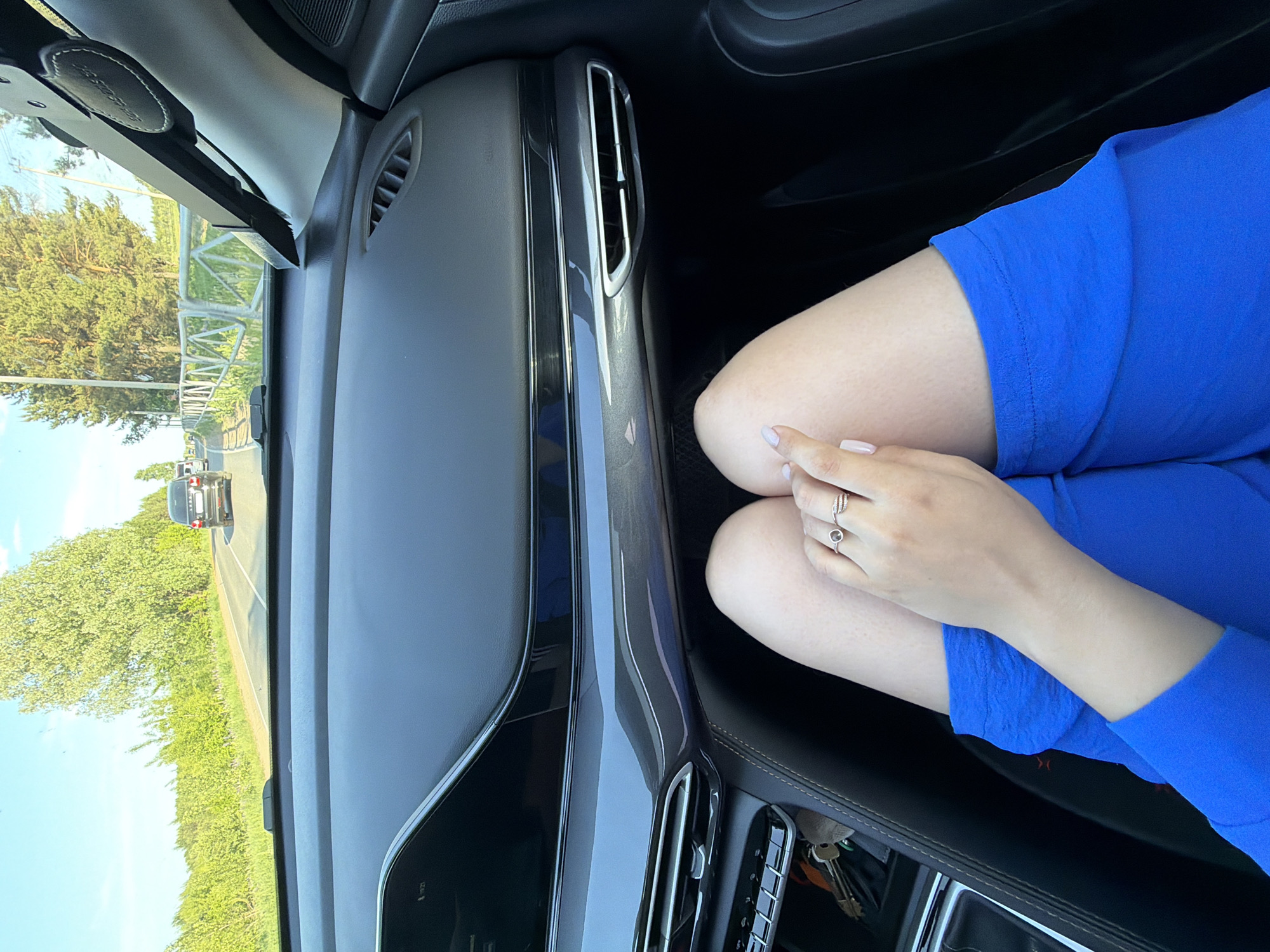 The scene depicts the interior of a car, potentially in motion, with the dashboard prominently displayed. Outside the windshield, a road and adjacent greenery are visible. The focus of the image is on a person's legs and arm, wearing a bright blue garment. The person has their hands clasped together, and a ring is visible on one of their fingers. Keys are present in the center console. There is no conclusive evidence to determine the location is Chaikovsky, Russia. The image shows only the inside of the car, the road outside and a bit of greenery.