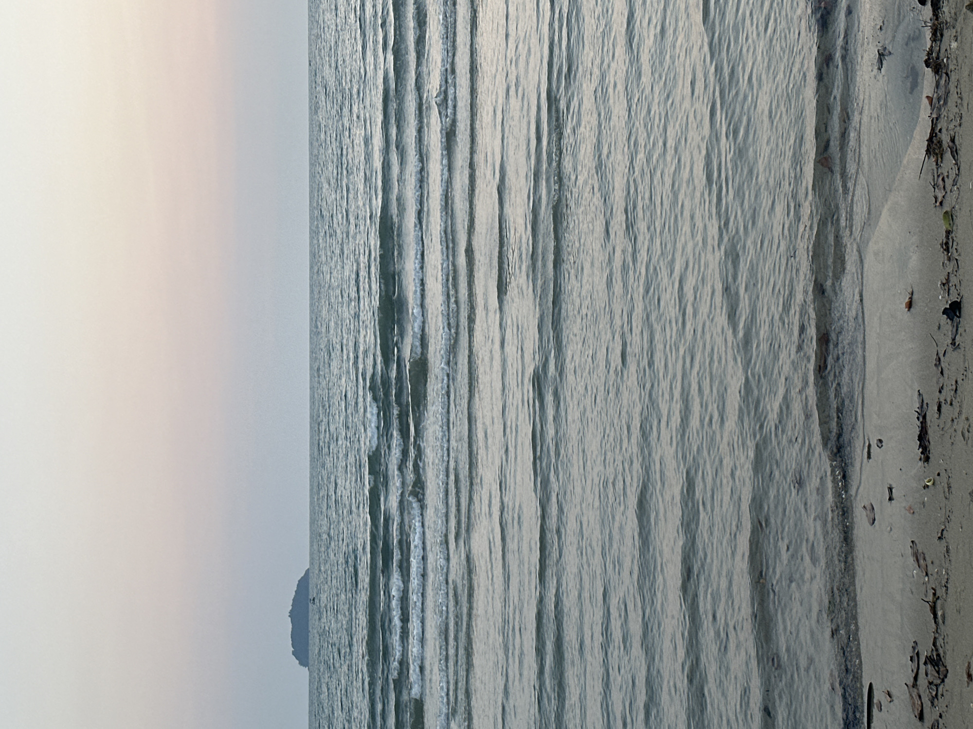 The image shows a view of the ocean and beach in Baan Klong Prao, Thailand. The beach is on the right side, transitioning to the ocean on the left. Gentle waves are visible throughout the water. The sky is pale, with a possible sunset or sunrise occurring. There is a landmass in the distance, partially obscured by the sky's haze. Debris litters the shoreline.