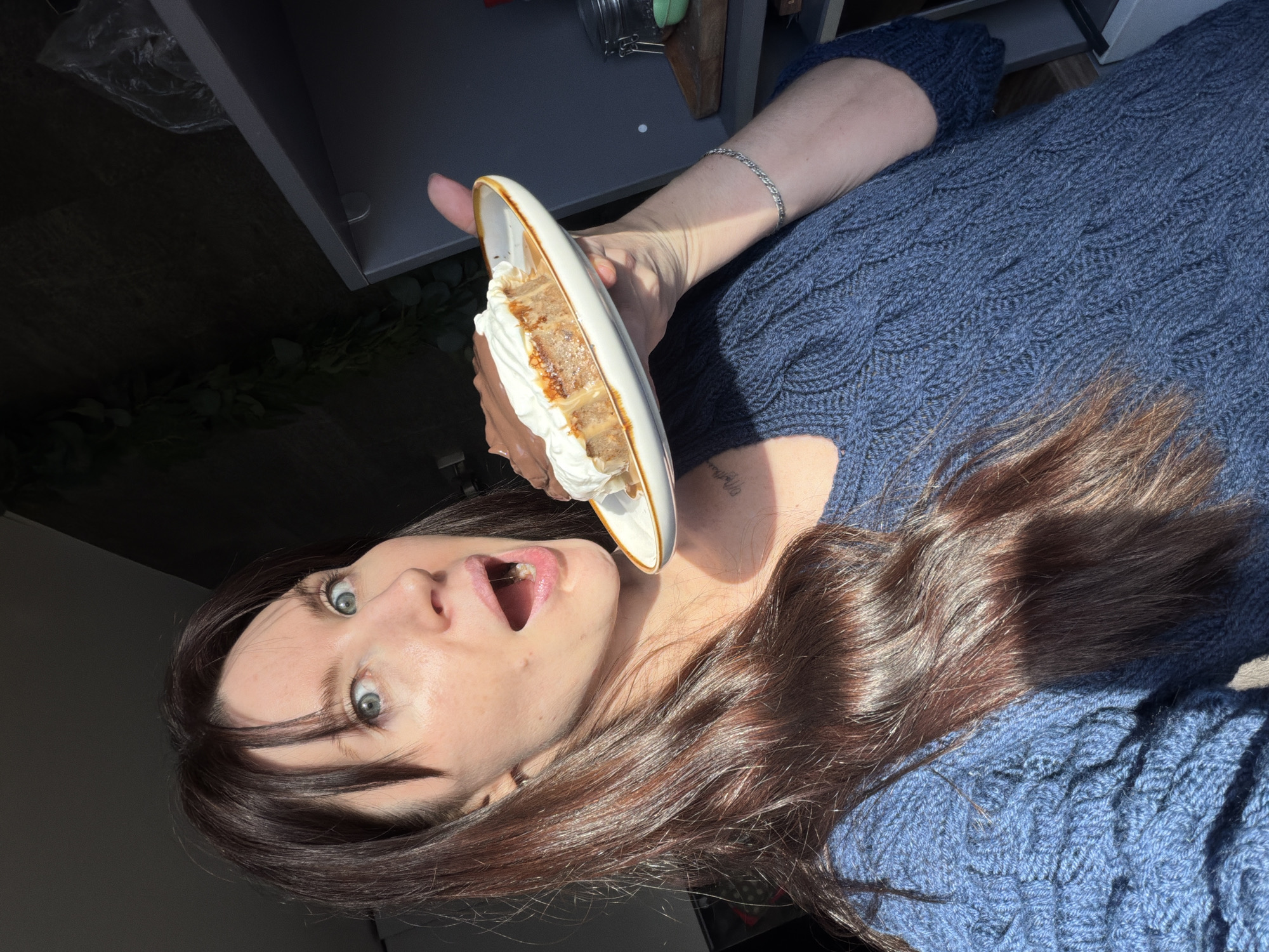 A medium shot, taken indoors, depicts a woman in a blue sweater holding a plate with a dessert. Her mouth is open in a gesture of apparent surprise or excitement. Her long brown hair is draped over her sweater, and her eyes are wide. Her left arm, adorned with a silver bracelet, holds the plate towards her mouth. The plate contains a slice of cake or dessert, topped with whipped cream and possibly a caramel drizzle. The background includes a dark-colored cabinet or shelf area, and there's a glimpse of what appears to be greenery. No definitive markers indicate the specific location of Perm, Russia. The image focuses on the woman's reaction and the presented dessert.