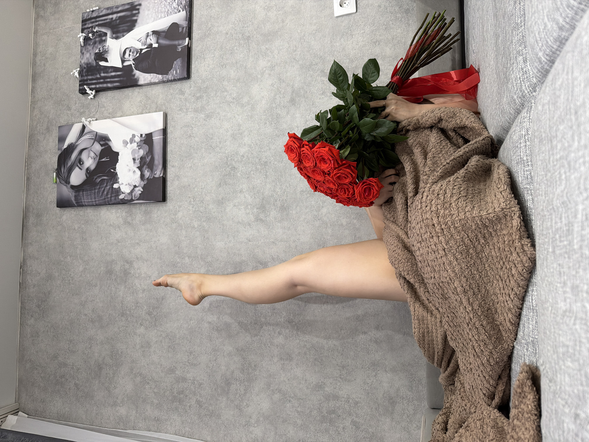 A person is lying on a gray couch against a gray wall in a room. The person is covered with a brown textured blanket and holding a large bouquet of red roses with a red ribbon. One bare leg is extended towards the wall. Two framed black and white photographs hang on the wall above the couch. A white electrical outlet is also visible on the wall. The photographs appear to be wedding portraits. The location is not explicitly specified but it could be Chaikovsky, Russia.