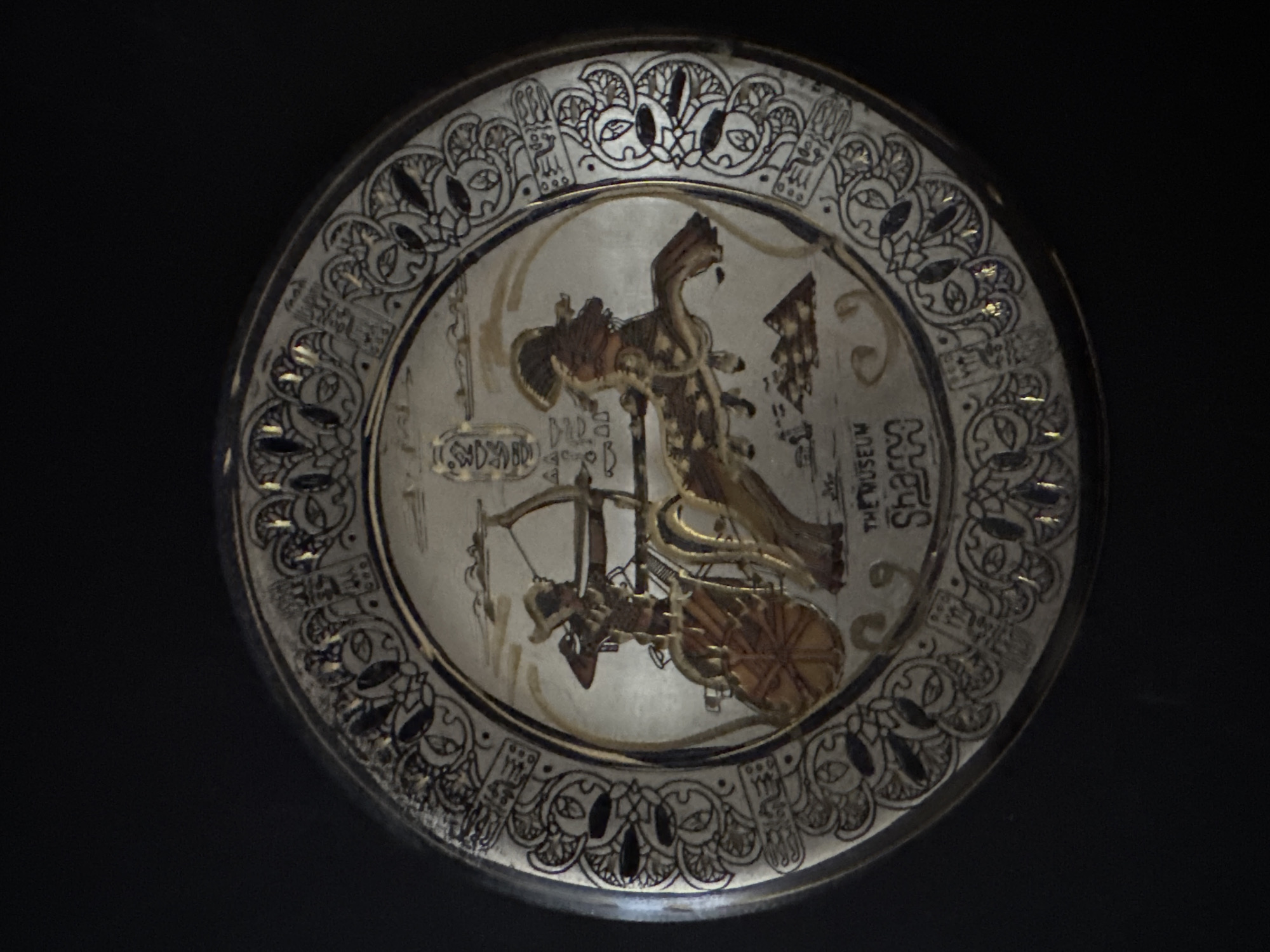 The image shows a decorative plate with an Egyptian-themed design. The plate is circular and has a metallic sheen, possibly silver or pewter. The central motif depicts an ancient Egyptian chariot with a warrior aiming a bow and arrow, pulled by a horse. Hieroglyphics are visible above the chariot and in the decorative border of the plate. The border is intricate, featuring stylized patterns and what appear to be lotus flowers. Text on the lower part of the plate reads "THE MUSEUM" and "Sharps". The plate is set against a plain black background. There is no indication of its location in Grodzisk Wielkopolski, Poland.