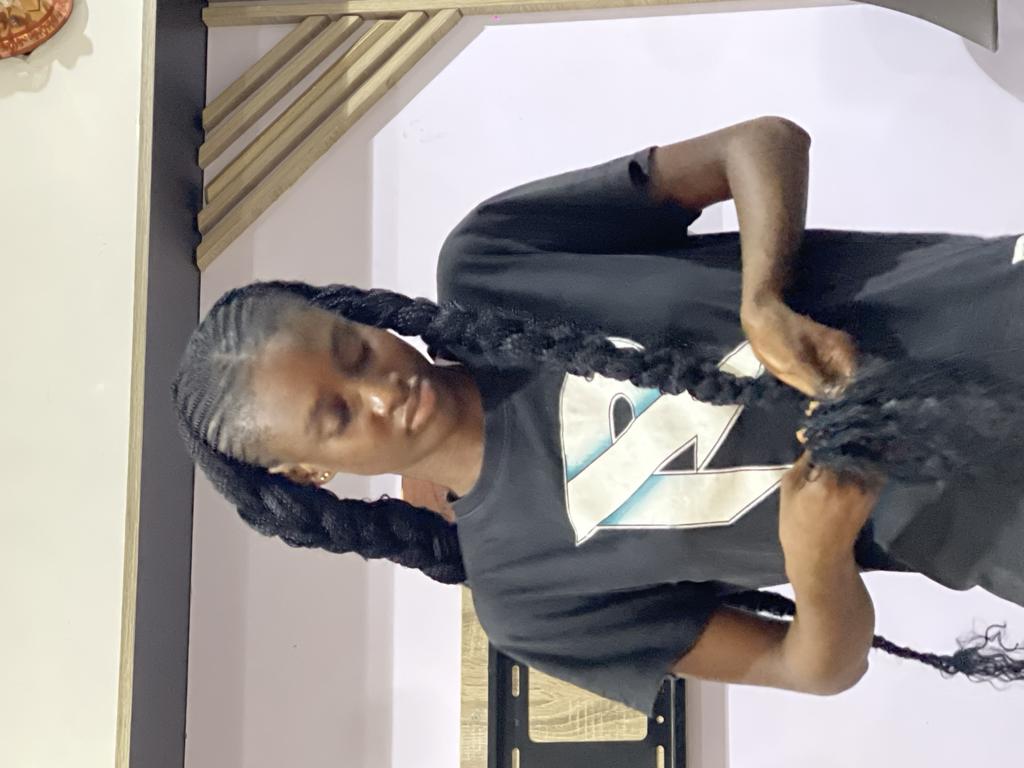 A young Black female is shown from the chest up, her dark hair styled into two large braids that hang down in front of her. She is wearing a black t-shirt with a graphic design on the front. Her hands are occupied with her hair, appearing to be in the process of braiding or adjusting it. The background is a plain wall with some wooden decorative elements on the right side. The lighting suggests an indoor setting, possibly a home. The location is identified as Abuja, Nigeria.