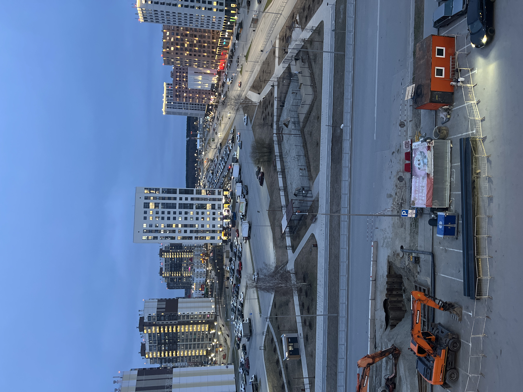 This image captures an urban scene in Tyumen, Russia, viewed from a high vantage point during dusk or early evening. The sky is a clear, deep blue, and numerous windows in the buildings are illuminated.
On the left side and in the background, a series of modern, high-rise residential buildings dominate the skyline, showcasing contemporary architecture with many glowing windows. Towards the center of the image, a wide, multi-lane street stretches into the distance, bordered by parking areas filled with numerous parked cars and commercial vehicles. Between the main road and the buildings, landscaped pedestrian zones with paths, benches, and small trees are visible.
In the foreground, on the right side of the image, there is an active construction site. Two orange Doosan excavators are positioned within a large excavated trench. Nearby, construction materials, pipes, and metal temporary fences are scattered. A small, orange portable cabin, marked with the number "1," stands next to a parked car.
Visible text includes "FANCYROOM.RU" and partial Russian text "Постельное белье" (Bed linen) on a banner near the construction area, "DOOSAN" on the excavators, and a blue "P" sign indicating parking. No people are distinctly visible, but their presence is implied by the illuminated buildings and parked vehicles. The scene suggests a busy residential or mixed-use area undergoing development.