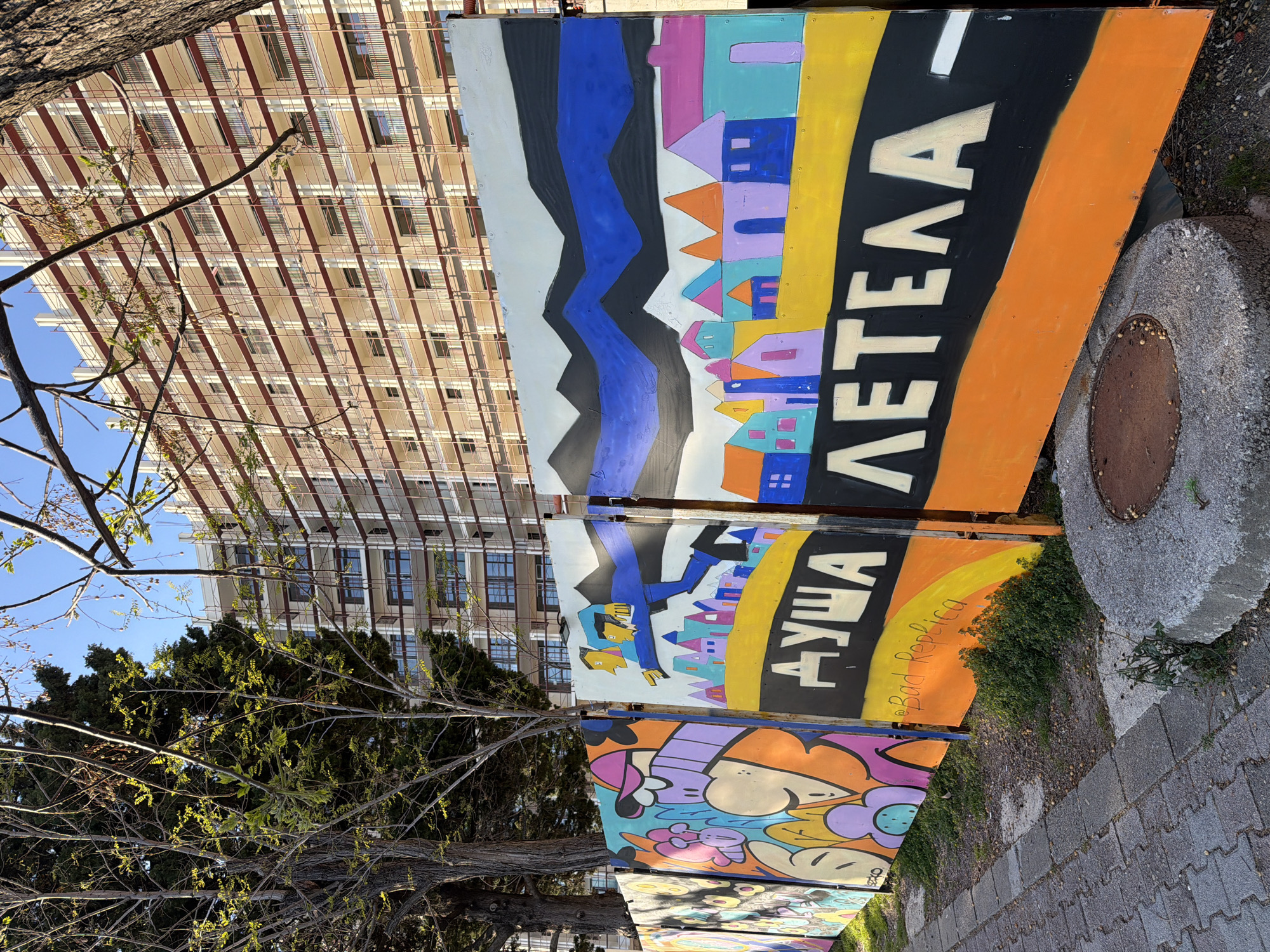 The image depicts a series of painted panels resting on the ground along a sidewalk in Rafailovići, Montenegro. The panels display colorful, cartoonish artwork. The top panel features an abstract depiction of mountains and houses. The second panel includes figures and text. A multi-story building is visible in the background. A tree is in the foreground. A sewer access point is in the lower right. The panels have some handwritten inscriptions and tag. The photo is taken from a low angle and rotated slightly.