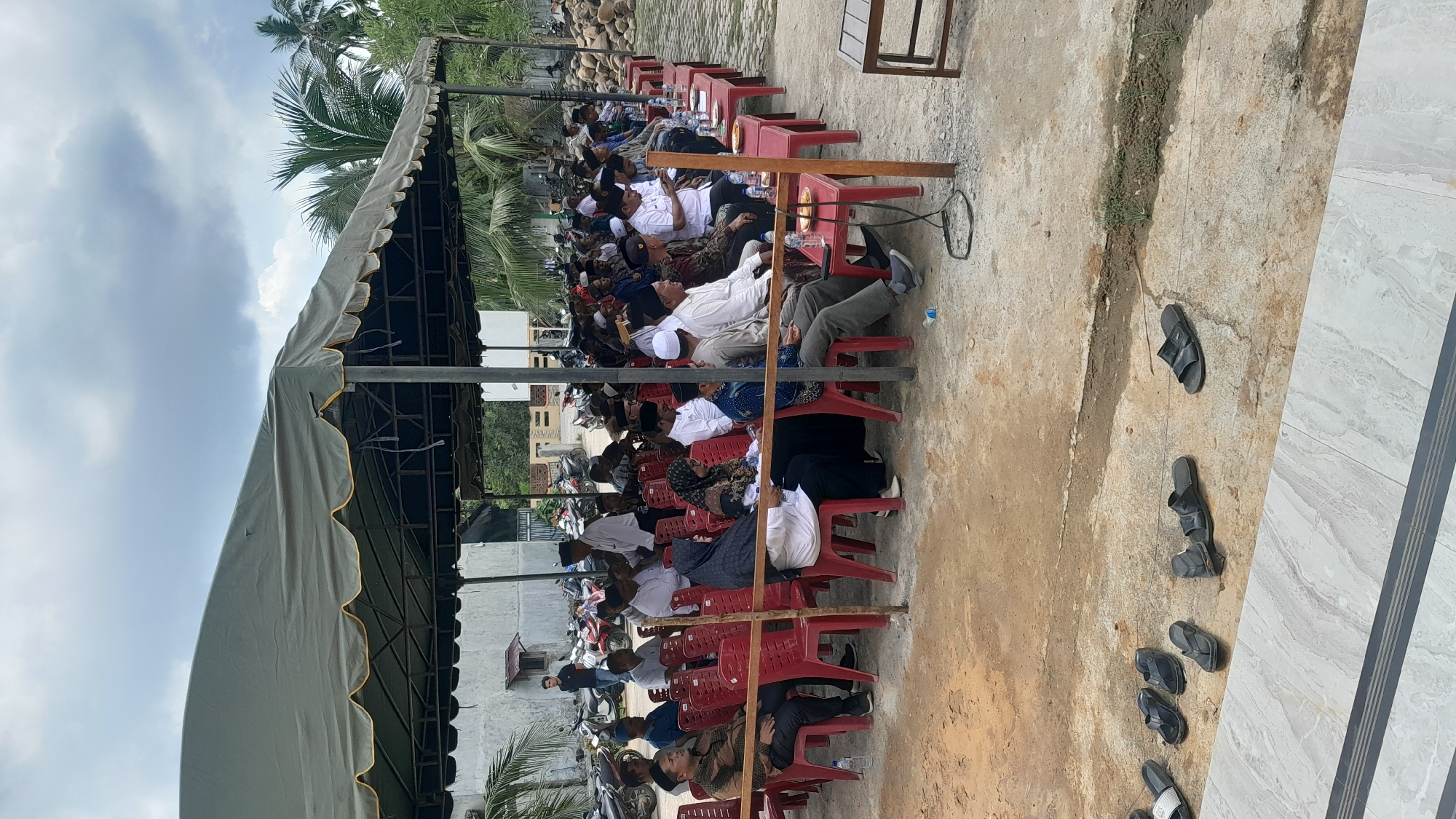 The image shows a gathering of people in Blangriek, Indonesia, seated on red plastic chairs under a large, tent-like structure with a grey canvas roof and metal supports. Most of the people appear to be men, some wearing white head coverings. They are seated in rows, with small tables between them. Behind the tent, there are trees, a building, and some parked motorcycles. To the right of the seated area, there are several pairs of sandals or shoes scattered on the ground, near a tiled surface. The sky is overcast.
