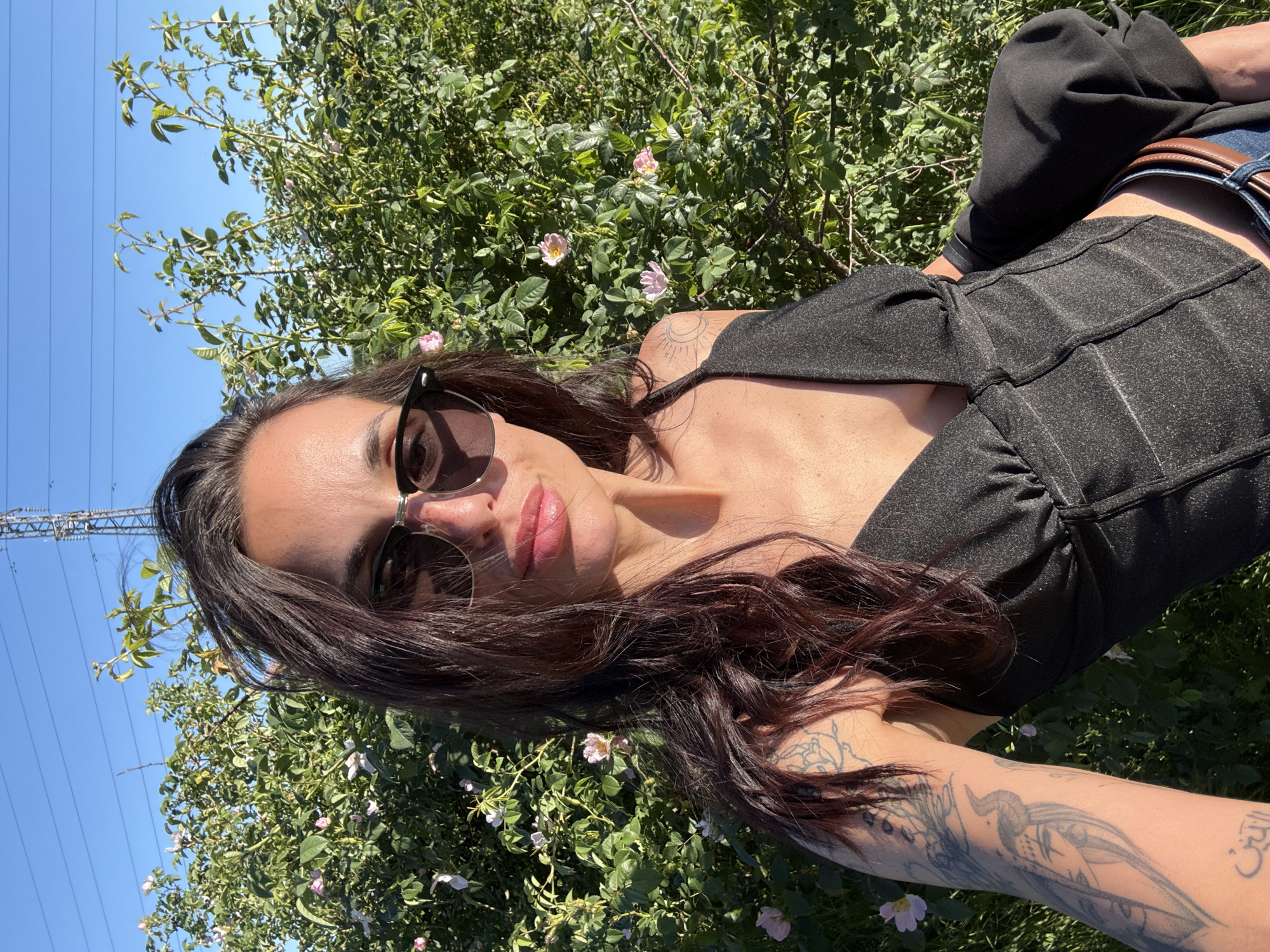 A woman is lying in a bush with pink flowers on a sunny day. She is wearing sunglasses and a black top. A tattoo is visible on her right arm. Utility poles with power lines are visible in the upper left of the image. Location is not specified within the image.