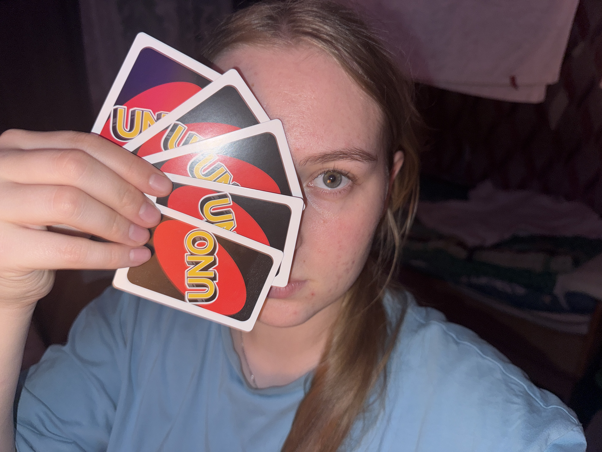 A woman with fair skin and light brown hair, partially obscured by several Uno cards, is holding the cards up to her face. The cards cover her nose and part of her cheek. She is wearing a light blue t-shirt. The background is indistinct, showing what appears to be bedding or clothing hanging on a dark wall. There is no indication of the location being Perm, Russia.