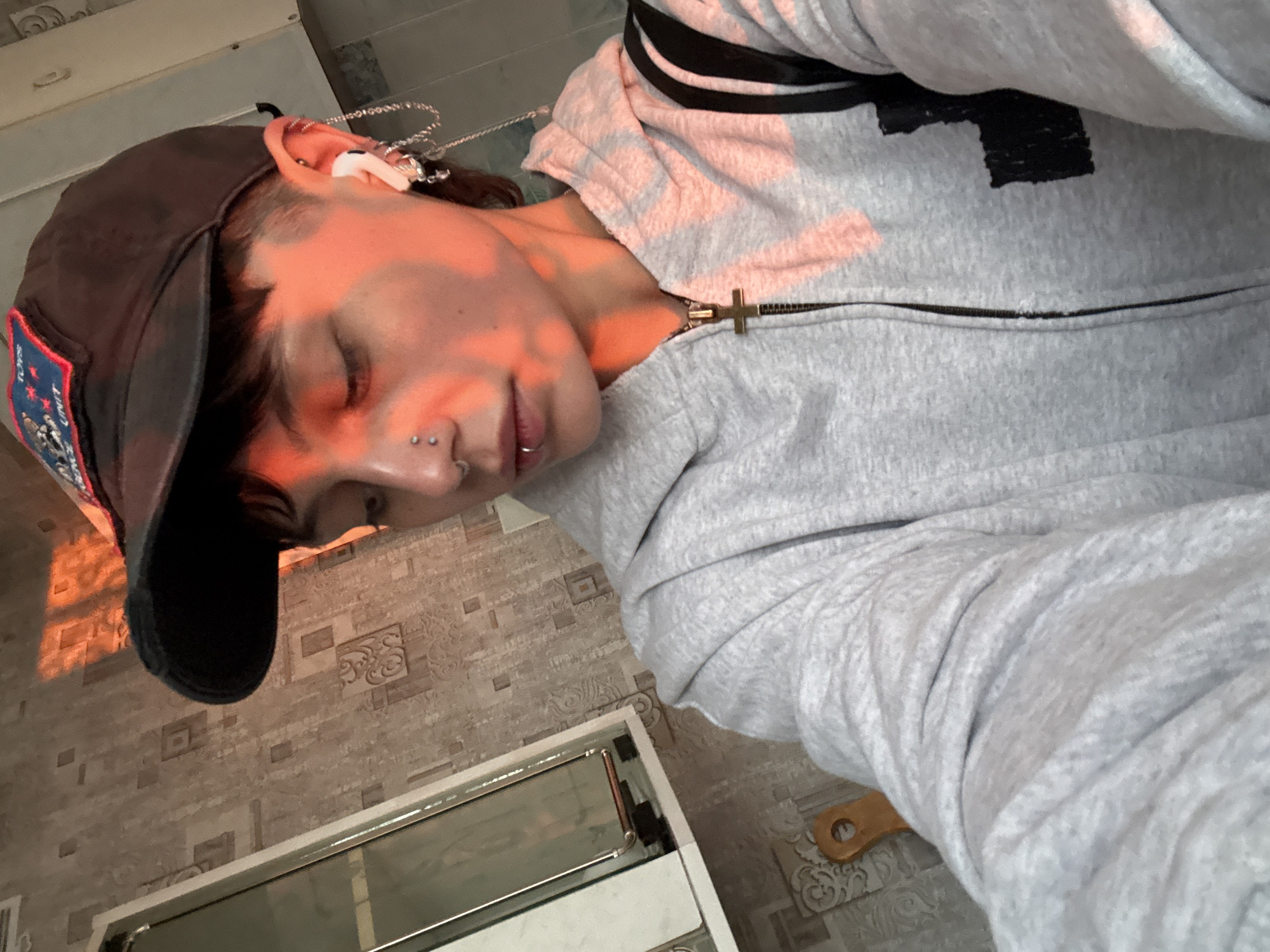 A person wearing a gray sweatshirt with black stripes and a black baseball cap is depicted in a close-up, eye-level shot. The person's face is illuminated by warm, orange-toned light, possibly sunlight filtering through a window, that emphasizes the nose, cheeks, and brow. The individual appears to have facial piercings on the nose and lip. Part of what looks like a white electronic device, possibly an earbud, is visible in the ear. A silver chain adorns the ear. The setting includes a tiled floor and part of a white object that looks like an oven. The surrounding environment is not clear enough to pinpoint the precise location in Saint Petersburg, Russia.