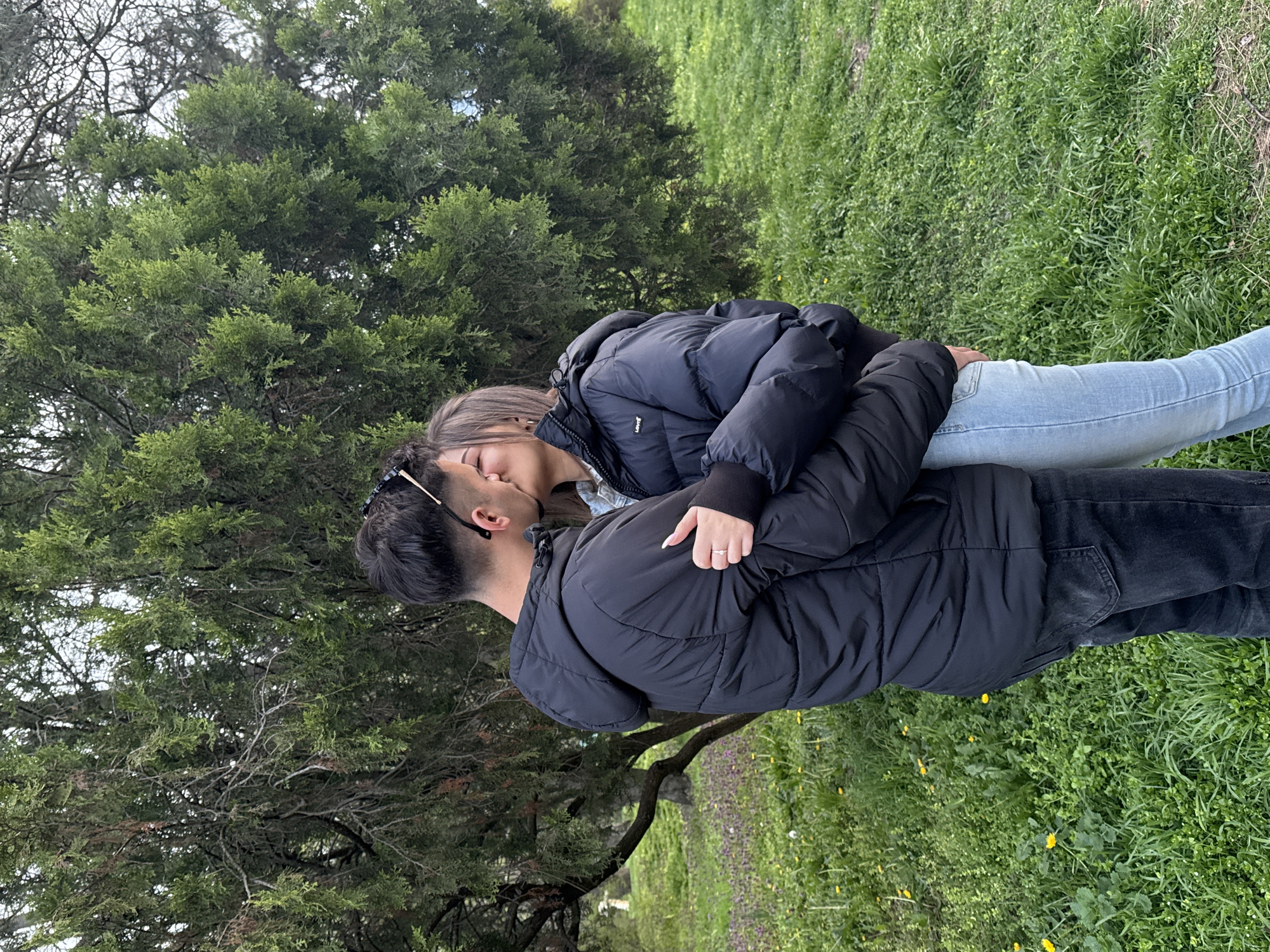 A photograph taken outdoors depicts a couple kissing in a park-like setting. The woman has medium-length brown hair and wears a black puffy jacket over a light-colored denim shirt and jeans. She has a ring on her left ring finger. The man, with short dark hair, wears a black puffy jacket and dark jeans. He also has sunglasses tucked into the back of his shirt. They are standing on a grassy area with trees in the background. Small yellow flowers are scattered throughout the grass. It is difficult to pinpoint the precise location in Sofia, Bulgaria, without additional landmarks.