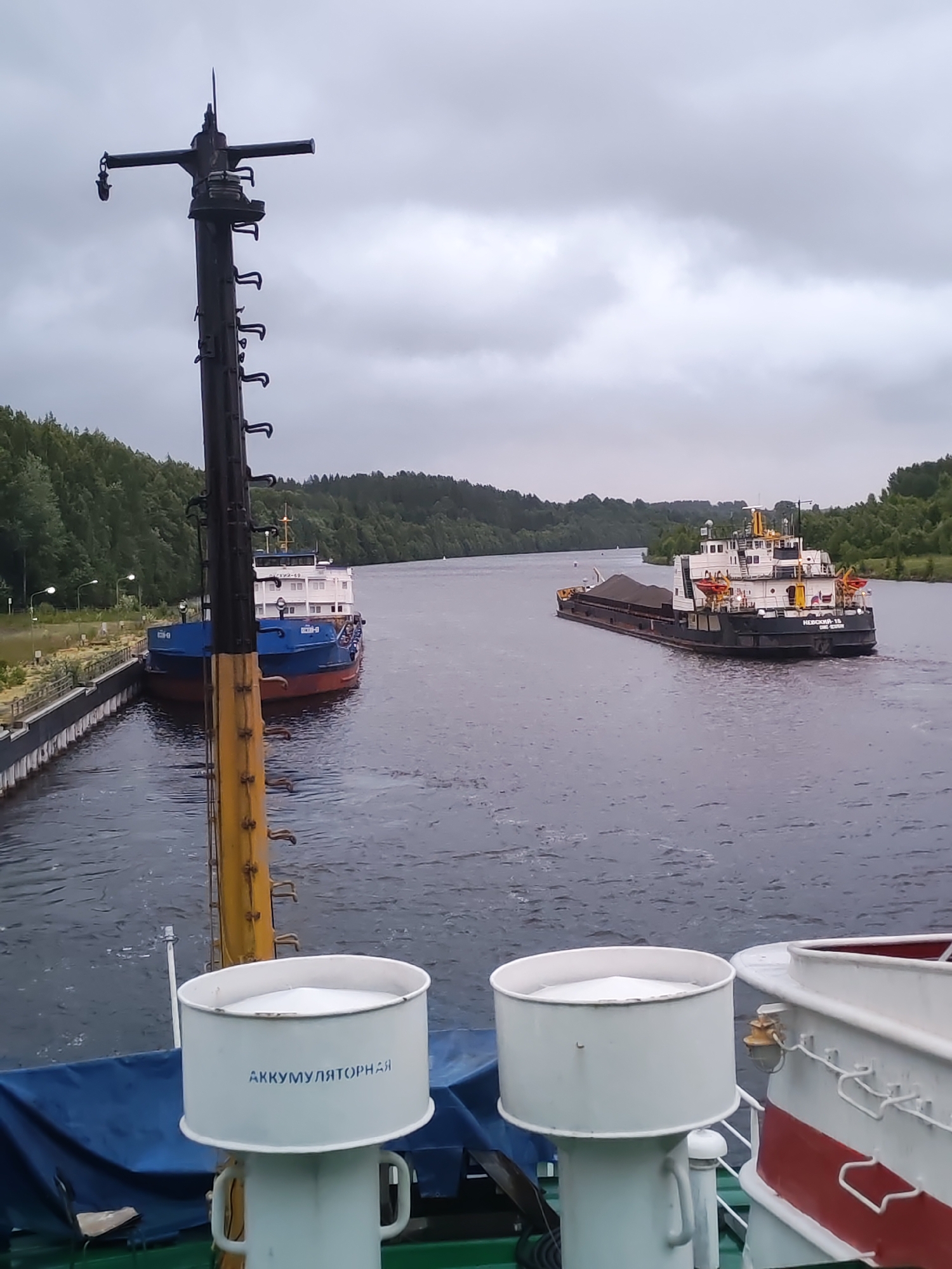 This image depicts a waterway with two vessels and a utility pole in the foreground. The waterway is surrounded by lush green trees on either side. The sky is overcast, with heavy clouds.
In the foreground, a tall, dark utility pole with crossbars and various attachments dominates the left side of the frame. Part of a vessel's deck is visible in the lower portion of the image, featuring two white, cylindrical containers with the Russian text "АККУМУЛЯТОРНАЯ" (Accumulator) printed on one of them.
On the left side of the waterway, a blue and red vessel is docked alongside a concrete structure with lights. On the right side of the waterway, a larger black and white vessel is moving away from the viewer, carrying a load of dark material in its cargo hold. This vessel has the name "НЕВСКИЙ-15" visible on its stern.
The location appears to be a canal or river. Given the presence of Russian text and the description of the scene, it is likely a canal or waterway within Russia. Alekseevskoe is a town in the Republic of Tatarstan, Russia, known for its woodworking industry and its location on the Kama River, which is part of the Volga-Kama waterway system. This waterway is a major transport route, consistent with the presence of cargo vessels. The scene suggests a functional waterway with ongoing shipping activity.