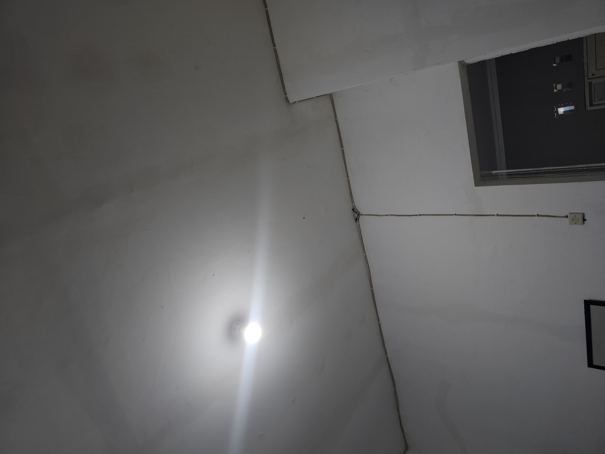 The image shows an interior scene with a lightbulb illuminating a white ceiling. Electrical wires are visible running along the ceiling and down a wall. A doorway to another room is also visible, with a glimpse of furniture and windows inside. The scene appears to be located in a residential or commercial building. The specific location in Lamongan, Indonesia cannot be determined from the image alone.