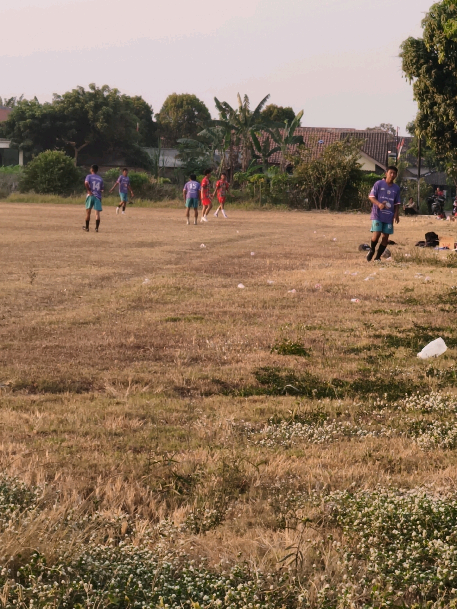 In Desa Sindon, Indonesia, a group of young men are playing soccer on a dry, grassy field. The field is littered with white plastic bottles and other debris. The sky is overcast with a pale pink hue. In the background, there are trees, houses, and some bystanders observing the game. The players are wearing casual athletic attire, mostly jerseys and shorts. One player in the foreground is wearing a purple jersey with the number 18 and turquoise shorts, and is facing the camera with a slight smile. Another player in a purple jersey with number 15 is visible on the right side of the field. A player in a purple jersey with number 14 is in the middle left. Two players in red jerseys are visible on the right side of the middle ground. The general atmosphere suggests an informal game in a rural or suburban setting.