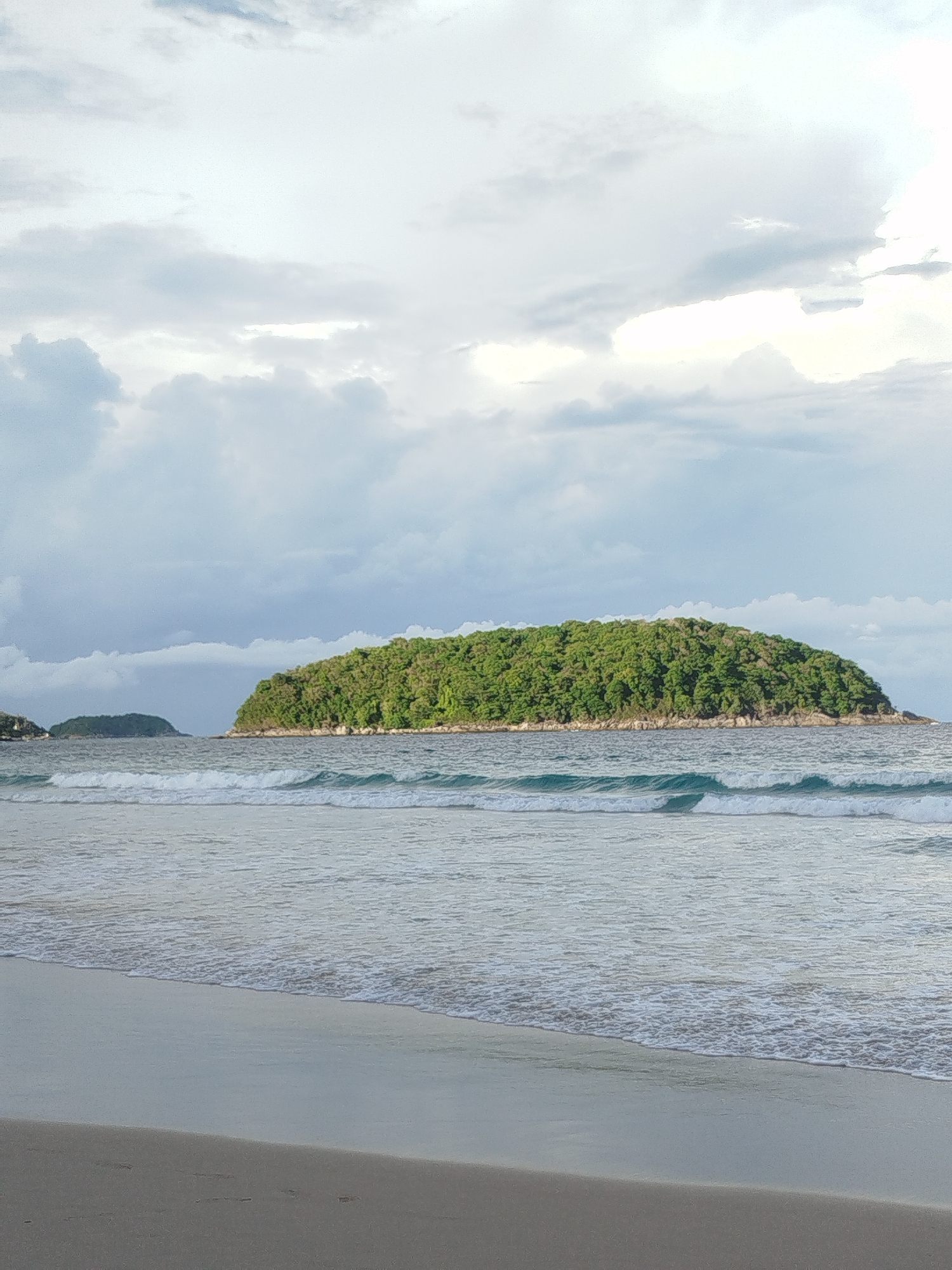 This is a full-frame, eye-level, vertical shot of a tropical beach with a lush, green island in the background, located in Rawai, Thailand. The foreground features a sandy beach with faint footprints and gentle waves lapping at the shore. The ocean is a calm, muted blue-green with white crests on the rolling waves. Beyond the water, a densely vegetated island rises from the sea, its slopes covered in a variety of green foliage. To the left of the main island, a smaller, similarly vegetated island is visible in the distance. The sky is overcast with a mix of light and dark grey clouds, with patches of bright white suggesting breaks in the cloud cover. The overall lighting is soft and diffused, characteristic of an overcast day. There are no people or animals visible in the image.