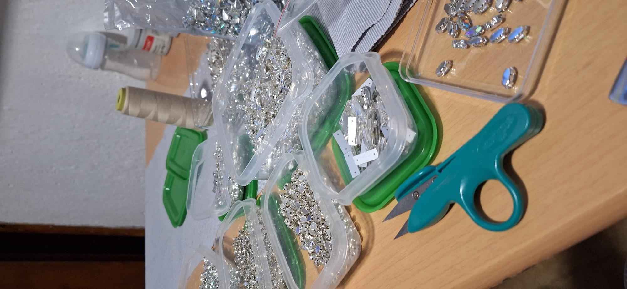 The image presents a close-up, overhead view of a crafting workspace, likely a table or desk, in an indoor setting. The primary subjects are numerous clear plastic containers, each filled with various types of sparkling rhinestones or crystals, suggesting preparation for an embellishment project.
The containers hold different shapes and sizes of rhinestones: some are small, faceted, and round, others are elongated rectangular pieces, and several are oval-shaped, with some exhibiting an iridescent or "Aurora Borealis" coating. Many appear to be sew-on rhinestones, featuring visible holes for attachment. The containers themselves are primarily clear plastic, some resting on or having green plastic bases/lids.
Among the crafting supplies, a large spool of beige or off-white thread with a light yellow core is visible to the left. A pair of small, teal-colored thread snips or scissors lies on the wooden surface towards the right side of the frame. Partially visible in the top left corner is a baby bottle with an unreadable red and white label. A light grey, textured fabric, possibly denim or a similar material, can be seen underneath some of the containers, further implying a textile-related craft.
The arrangement of materials indicates an active or impending crafting session, likely involving sewing or attaching these decorative rhinestones to fabric. The lighting suggests an indoor environment. No specific time of day or emotions are discernible. While there is faint text on the baby bottle and possibly a logo on the snips, it is not legible. The given location context for this activity is Blagoevgrad, Bulgaria.