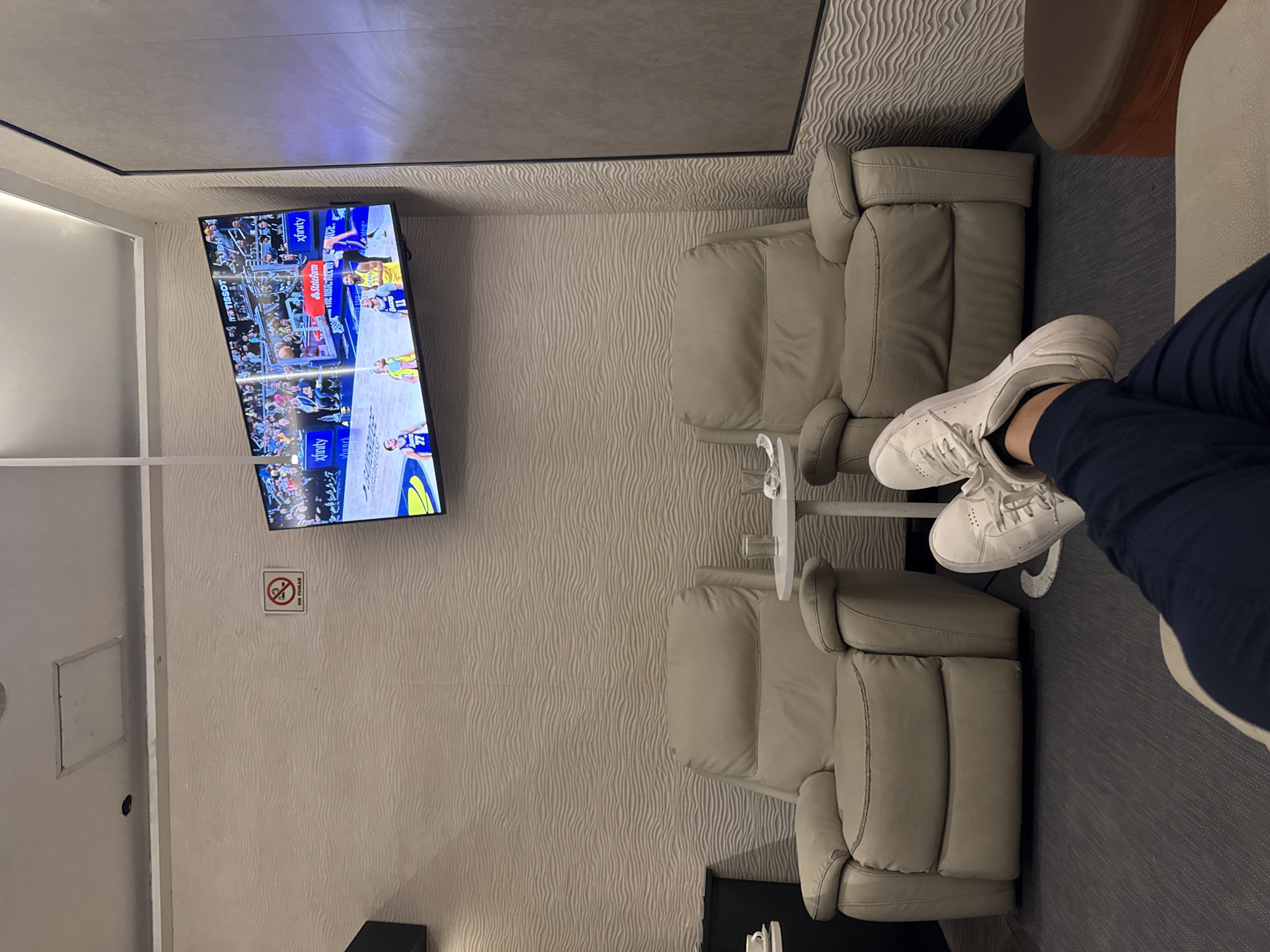 The image shows a room with two light gray recliner chairs facing a wall-mounted television displaying a basketball game. There is a small white table between the chairs. A person wearing blue pants and white sneakers has their legs crossed on the right side of the image. The walls have a textured, wavy pattern. A "No Smoking" sign is visible on the wall near the TV. The location is identified as being in Mexico City, Mexico.