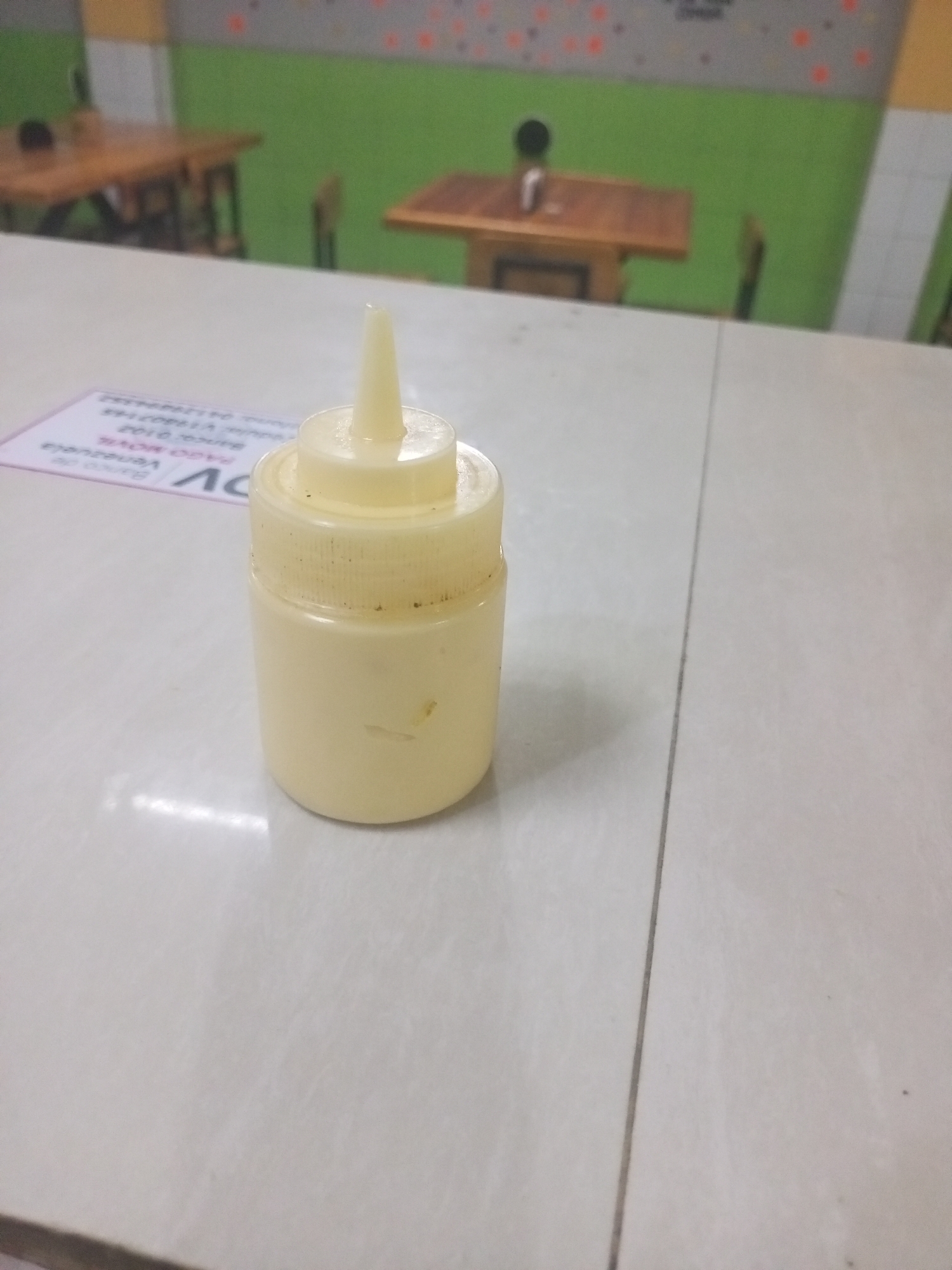 A plastic condiment bottle, likely containing mayonnaise or a similar sauce, sits on a light-colored tiled table. The bottle has a pointed nozzle for dispensing the contents. In the background, wooden tables and chairs are visible against a green and grey wall. A sign with text, possibly "Banco de Venezuela" and "Juan Griego," is partially visible to the left of the bottle, suggesting the location is Juan Griego, Venezuela.