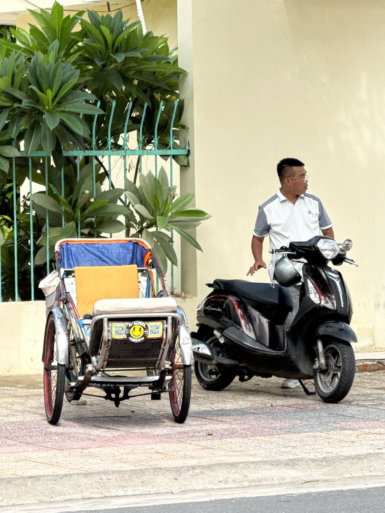 A man wearing a white and gray polo shirt and glasses stands next to a black scooter on a sidewalk in Nha Trang, Vietnam. He is facing to his right, looking away from the camera. To the left of the scooter is a three-wheeled pedicab with a blue canopy and an orange cushion. The pedicab has stickers on the back, including one with a smiley face and text. Behind the vehicles is a teal metal fence with lush green foliage. The building in the background has a pale yellow wall. The sidewalk is paved with stone tiles.