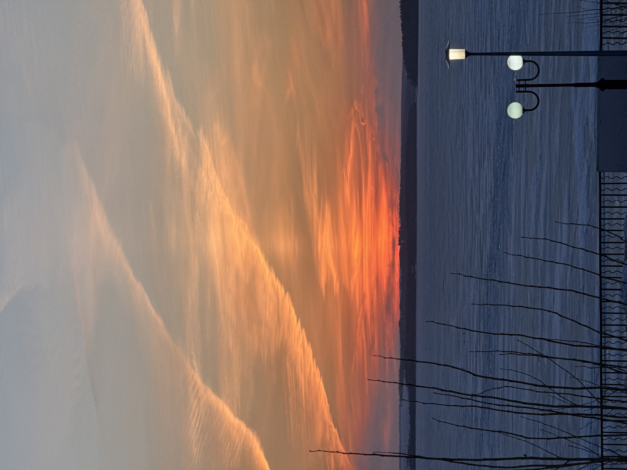 The image depicts a twilight scene in Votkinsk, Russia, with a sunset casting orange and yellow hues over a large body of water, possibly the Votkinsk Reservoir. A dark strip of land, likely the opposite shore, is visible on the horizon. In the foreground, a row of lampposts with spherical lamps illuminates a dark embankment, with bare tree branches silhouetted against the water and sky. The scene is calm and still, capturing the transition from day to night over the reservoir.