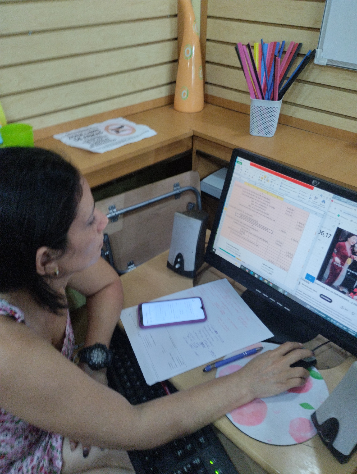 This image depicts an indoor office or workspace located in Juan Griego, Venezuela. A female subject, with dark hair, is seated at a light wood-colored desk, viewed from her left side. She wears a floral-patterned top and a dark-colored watch on her left wrist. Her right hand operates a black computer mouse situated on a circular white mousepad decorated with pink peach designs and green leaves.
A black computer monitor is positioned on the desk, displaying a spreadsheet application with numerical data on the left half and a video or image featuring a woman in a red dress on the right half. To the left of the monitor, a purple smartphone rests on a stack of white papers containing handwritten notes, with a blue pen placed on top. A silver computer speaker is located next to the papers.
The background consists of light-colored, horizontally slatted wall panels. Above the desk, a higher shelf holds a tall, orange conical decorative object with green circular patterns and a white perforated container filled with assorted colored pens or straws (including pink, black, blue, yellow, and red). A white paper displaying a "no firearms" symbol (a handgun with a red circle and slash) is visible on the left side of this shelf, and a portion of a white board is visible on the right. A green cup is partially visible on the far left of the desk area. The subject is actively engaging with the computer and its peripherals.