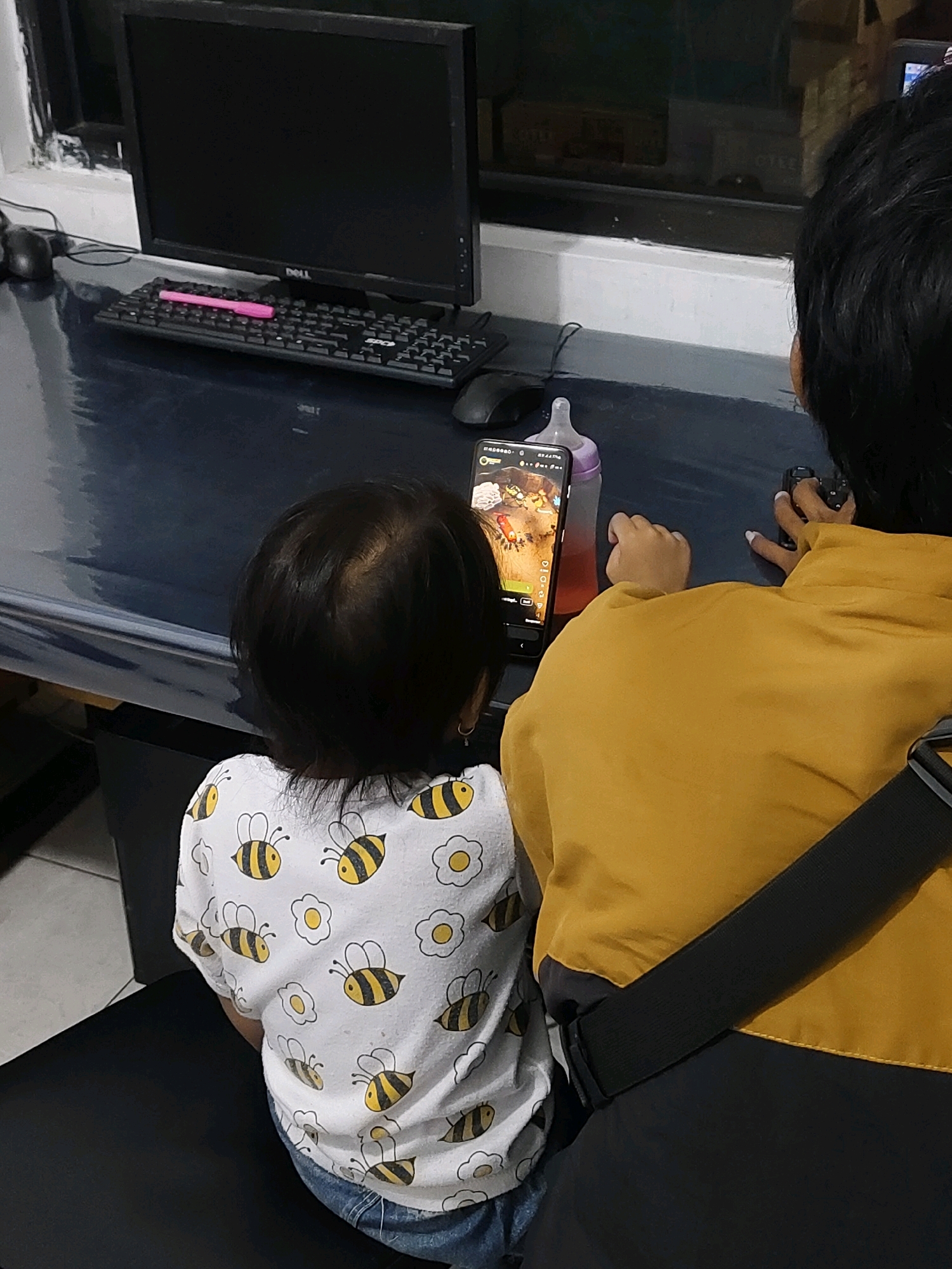 In Probolinggo, Indonesia, an indoor scene depicts a desk with a computer monitor and keyboard. A young child, viewed from behind, wears a white t-shirt with a bee and flower pattern and blue jeans. The child is looking at a smartphone displayed on the desk. To the right, an adult, also viewed from behind, wears a mustard yellow jacket with a black strap across the chest. The adult is holding a small camera in their right hand. A baby bottle is placed next to the smartphone. The overall setting appears to be a home environment.