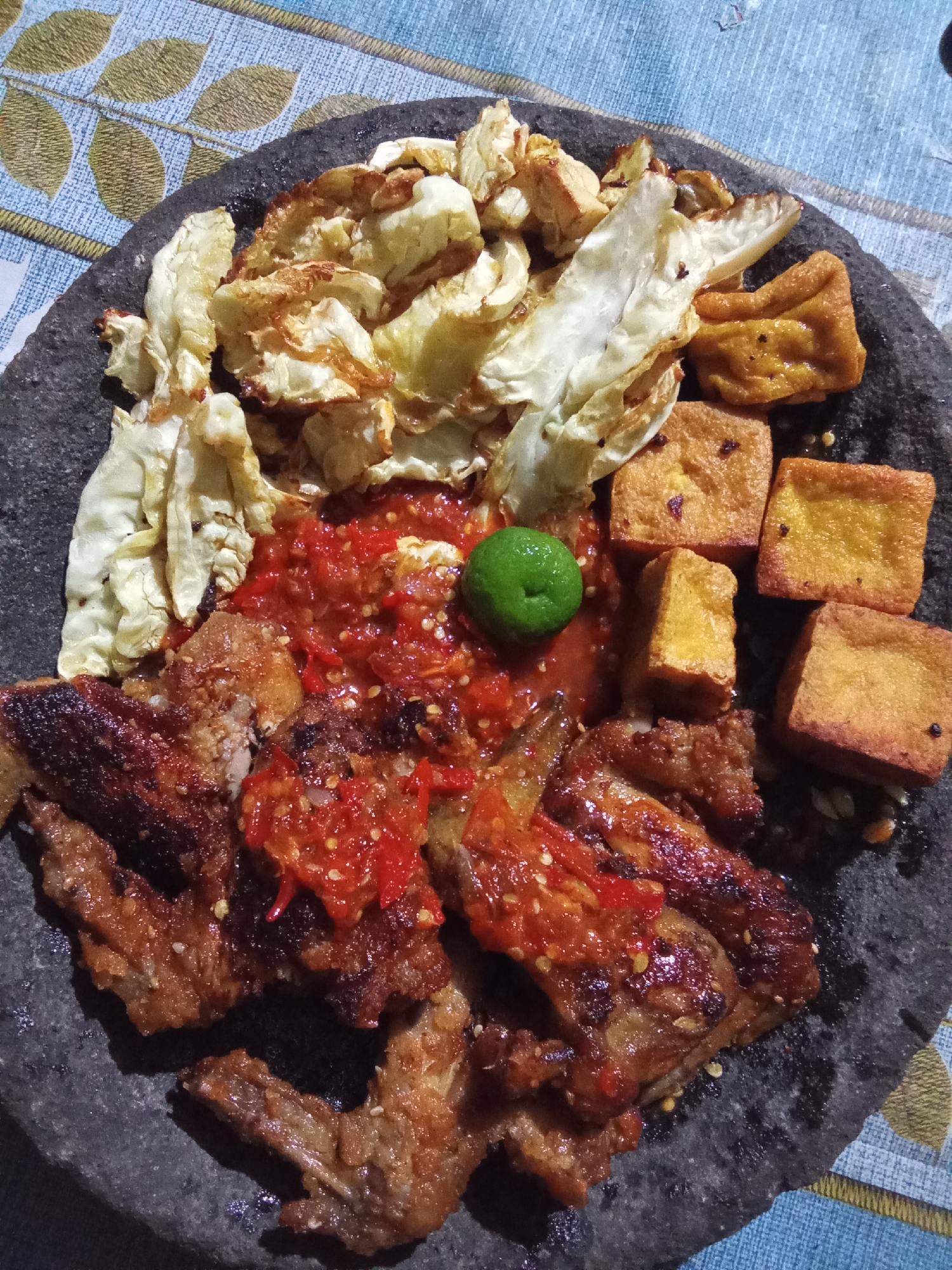 The image depicts a plate of food on a patterned tablecloth. The food is served on a dark, rough-textured stone slab and includes several items:
* **Fried chicken:** Several pieces of fried chicken, appearing crispy and coated in a red sauce, are arranged on the left side of the slab.
* **Sambal:** A generous portion of red sambal (Indonesian chili paste) is spread over the chicken and in the center of the plate.
* **Fried tofu:** Several cubes of golden-brown fried tofu are placed on the right side of the slab.
* **Cooked cabbage:** Pieces of cooked cabbage, appearing slightly wilted and browned, are arranged at the top of the slab.
* **Lime:** A single small green lime is positioned within the sambal.
The tablecloth beneath the stone slab has a blue and white pattern with green and gold leaf motifs. The lighting suggests the photograph was taken indoors.
The user asked for the location to be in the city of Subang, Indonesia. However, the image itself does not provide any visual cues or context that would confirm this specific location. Therefore, it's impossible to verify or determine the location based solely on the provided image.