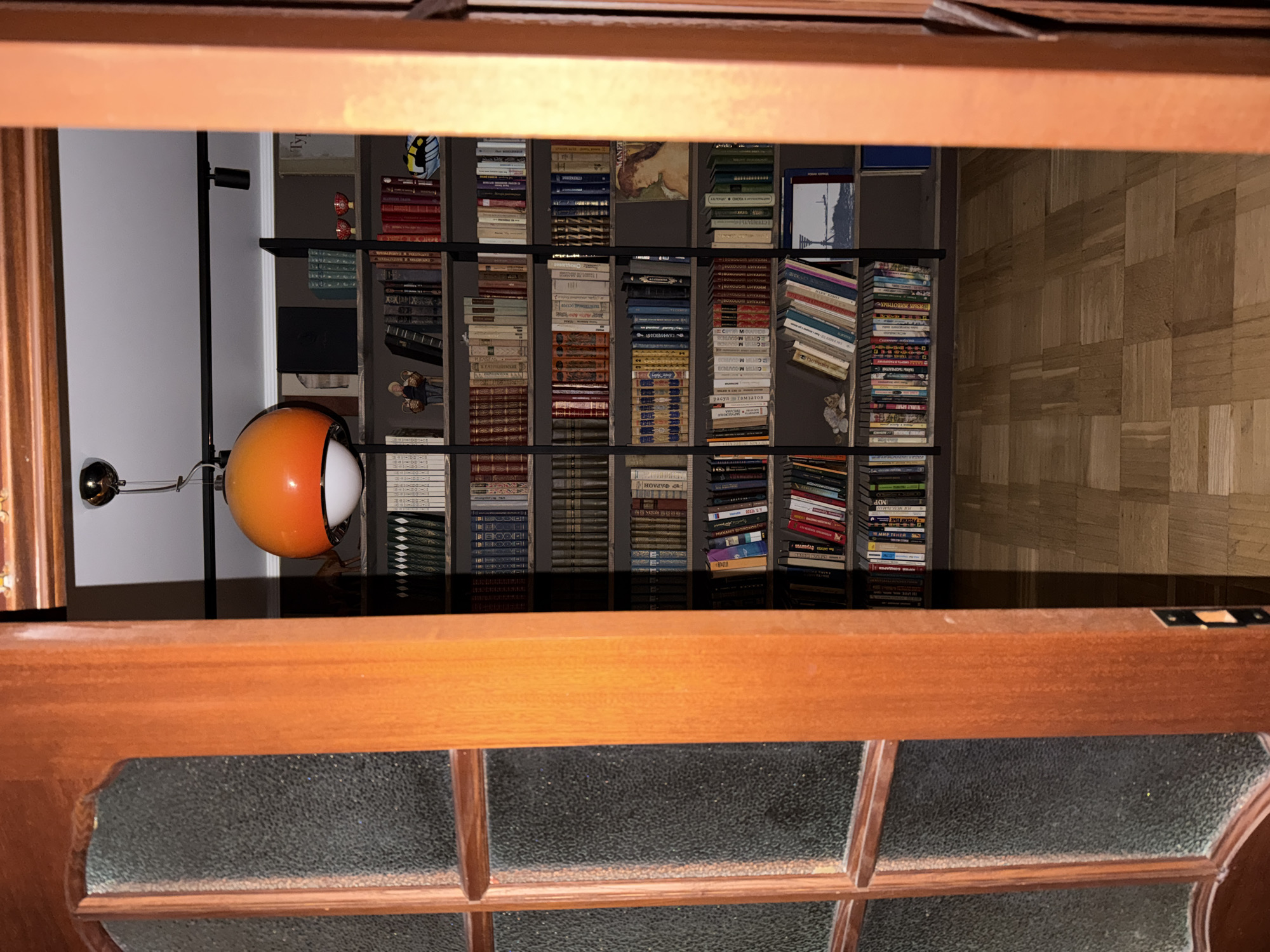 The image is an interior shot looking upwards through a partially open door, revealing a view of a bookshelf and a lamp. The bookshelf is filled with numerous books of various sizes and colors. A lamp with an orange and white globe is visible to the left of the bookshelf. The room has parquet flooring. The door is made of wood and has textured glass panels. The image does not contain any information about the location in Khimki, Russia or any people, subjects, or actions.