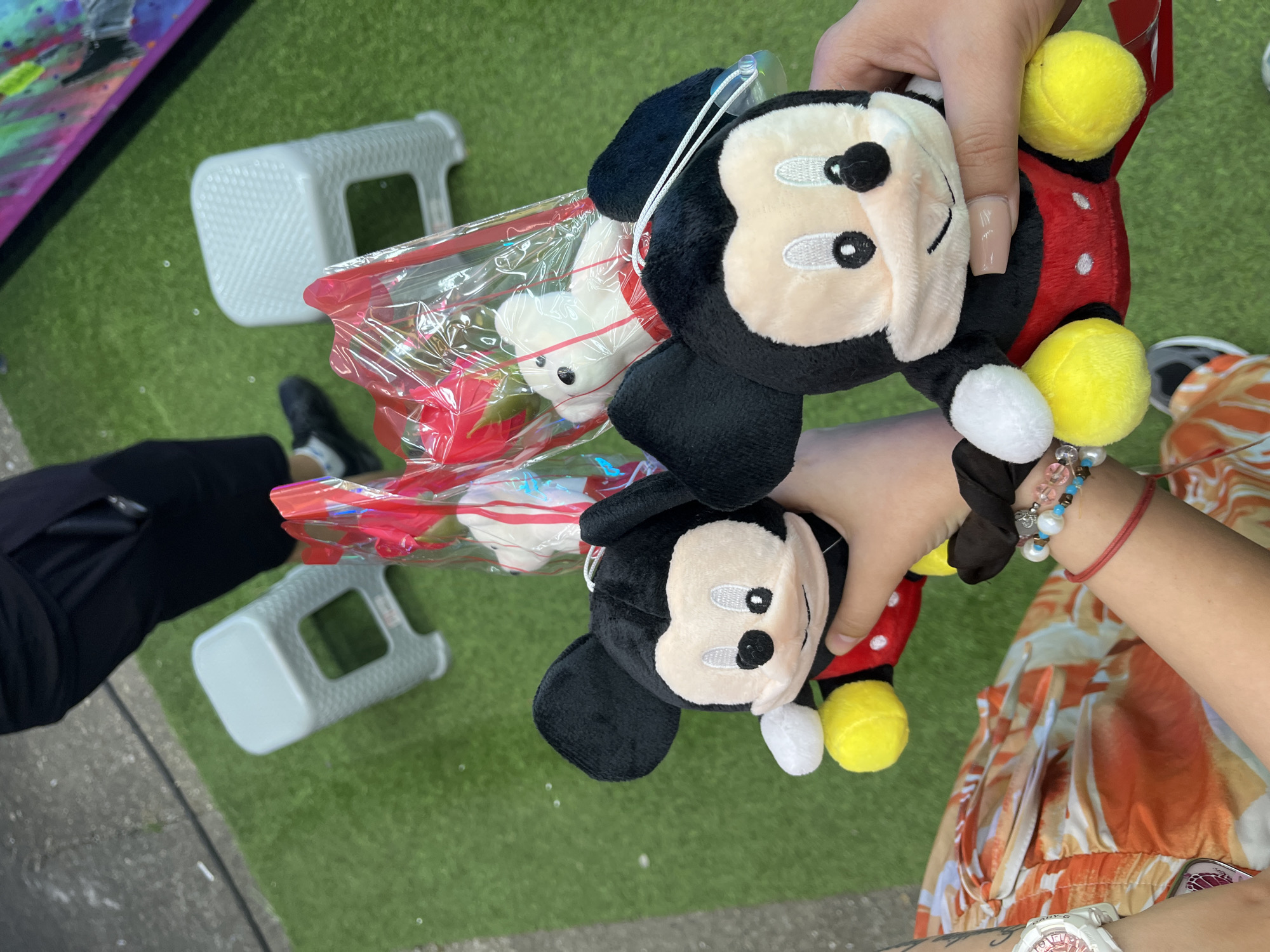 The image shows a close-up of two plush Mickey Mouse toys being held by two people. The toys are black and red with yellow shoes. In the background, there are two small white stools and a green artificial turf surface, suggesting an outdoor setting. A person's legs and lower torso, wearing black shorts and black socks with white shoes, are visible in the top left corner. Another person's arms and torso, wearing an orange and white patterned garment, are visible on the left side of the image. Bracelets are visible on one of the arms. The scene is not identifiable as being in Iskar, Bulgaria or any specific city.