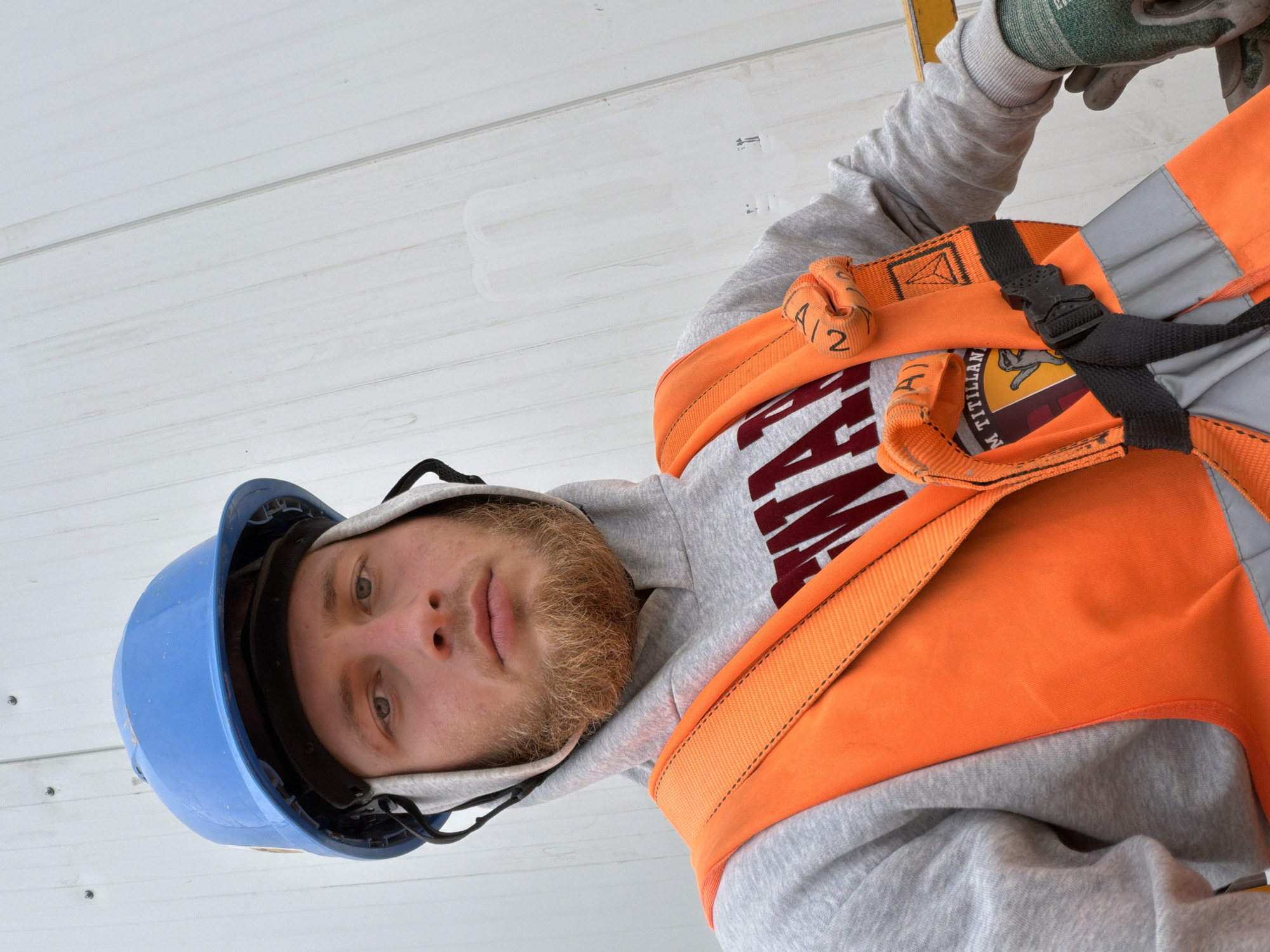 A young man with a ginger beard and blue hard hat is pictured against a white, corrugated wall. He is wearing a grey hoodie with the word "WAR" emblazoned on it in maroon letters, and an orange safety vest over the hoodie. The vest has black straps and reflective strips. His right hand, wearing a green glove, is visible at the lower right of the frame. The man appears to be at a construction site, possibly in Stróża, Poland, given the context provided.