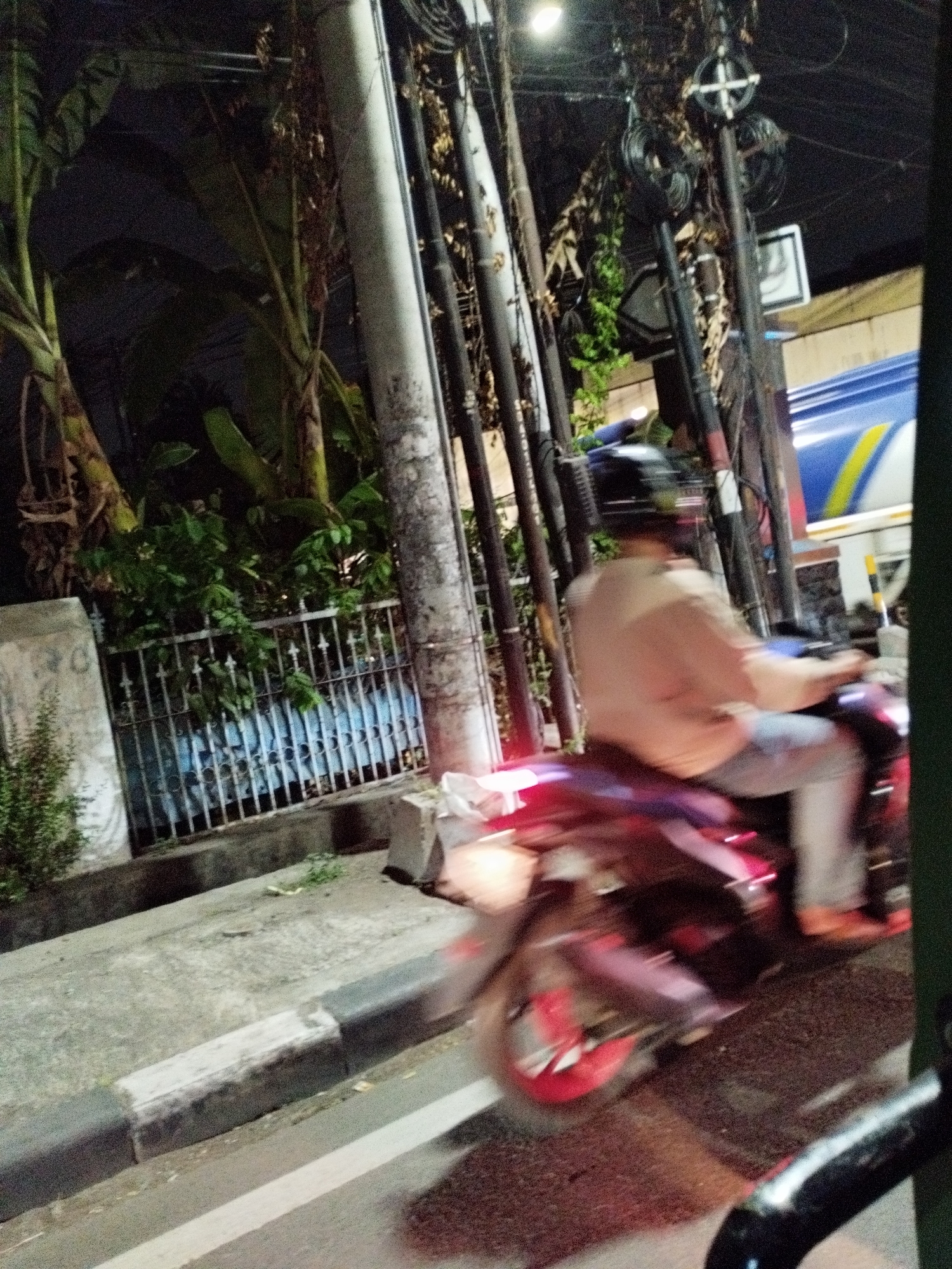 The image depicts a street scene at night in Surabaya, Indonesia. A person is riding a motorcycle, the movement of which is blurred due to motion. The rider is wearing a helmet and a light-colored shirt, with light blue jeans and orange shoes. The motorcycle itself is dark colored with red accents on the wheels. To the left of the motorcycle, a metal fence is visible, behind which are plants and what appears to be a blue wall. Several utility poles with numerous wires are present in the background, along with a portion of a large vehicle, possibly a truck or bus, with blue and yellow markings. The street is paved, with a curb that has white and black markings. The overall lighting suggests it is nighttime, with some streetlights and the motorcycle's taillight providing illumination.