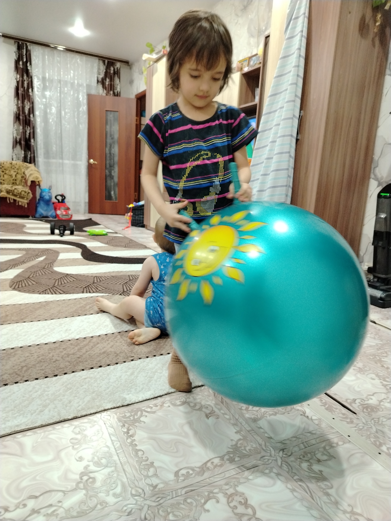 A young girl in a striped shirt stands next to a smaller child sitting on the floor, holding a large blue inflatable ball with a yellow sun design. They are indoors, likely in a home in Большая Уса, Russia, judging by the location information. The room has a layered rug with brown and white stripes and a patterned tile floor in the foreground. In the background, there is a brown door, curtains, and some toys. To the right, there is a piece of furniture and what appears to be a vacuum cleaner. The girl is standing and holding the ball, while the smaller child is seated on the floor next to her.