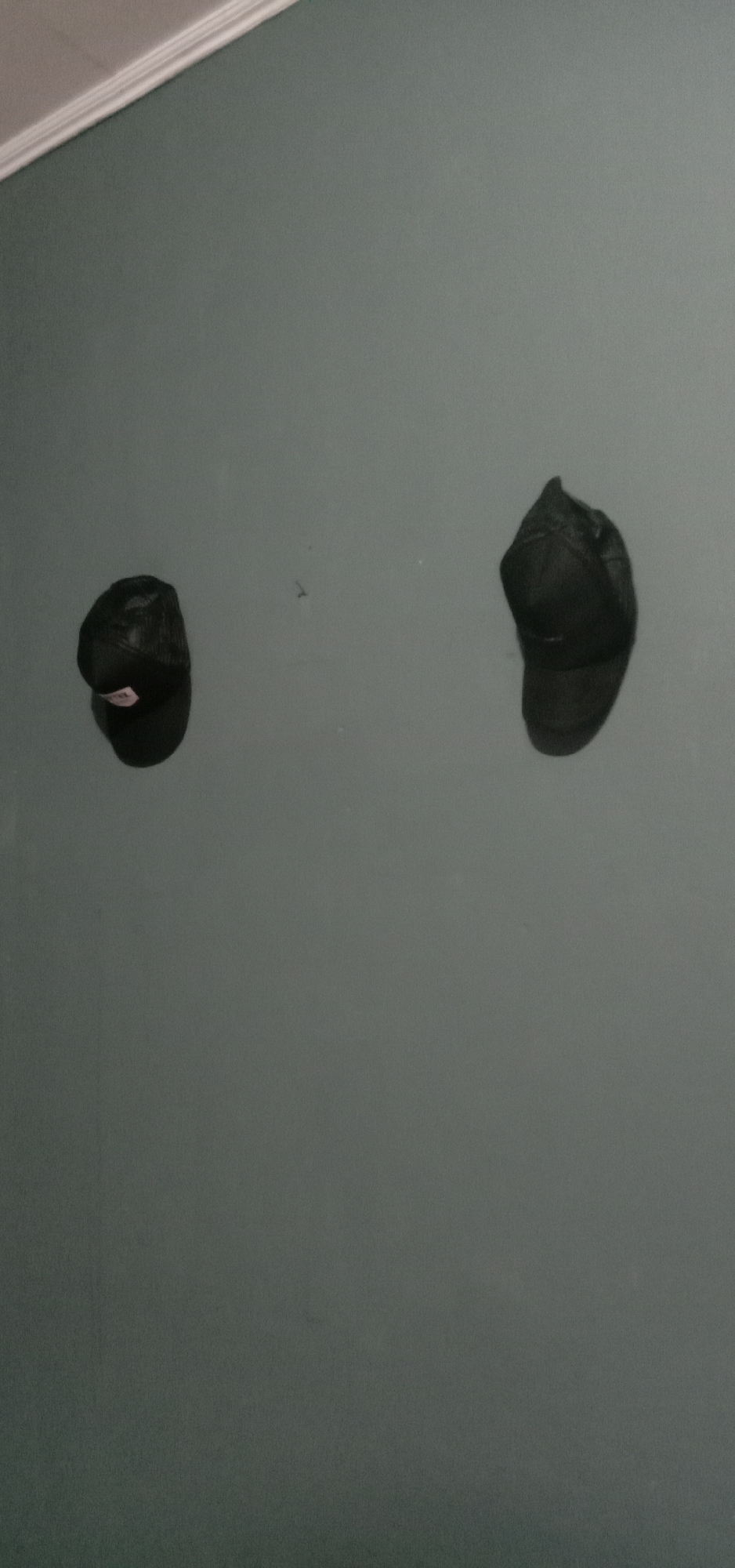 In an indoor setting, likely a room, two black baseball caps are affixed to a muted teal wall. One cap is positioned to the left, slightly lower than the other cap, which is located to the right. The caps appear to be made of a mesh material on the sides and a solid material on the front and top. The wall has a smooth finish and meets a white molding at the top edge of the frame. There are no people present, and the scene is static. The location in Kendari, Indonesia is not ascertainable from the visual information provided.