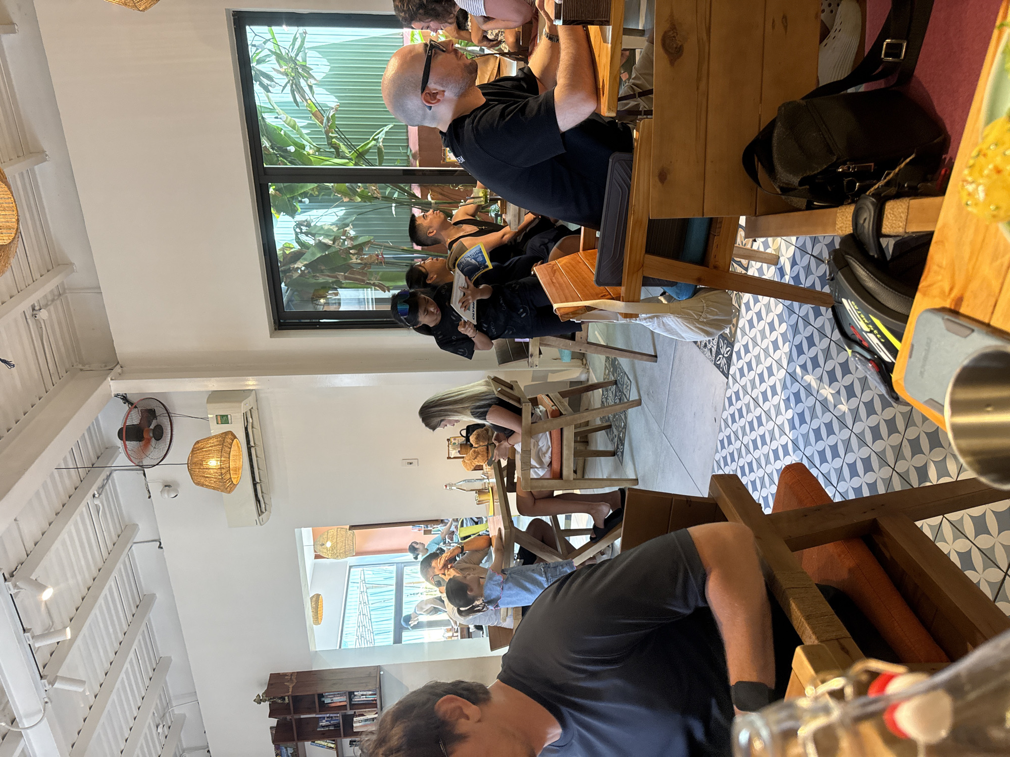 The image captures an interior scene of what appears to be a cafe or restaurant, filled with people seated at wooden tables. The setting is bright, illuminated by natural light streaming in through large windows. A variety of individuals occupy the space, with some engrossed in reading, while others are engaged in conversation.
The cafe features patterned floor tiles, pendant lighting, and a visible air conditioning unit and fan mounted on the ceiling. A bookshelf is visible in the background, suggesting a relaxed and intellectual atmosphere. People are wearing casual clothing, and some are seen holding books or electronic devices, suggesting a casual, informal setting. A few bags and personal items are also present on the tables and floor.