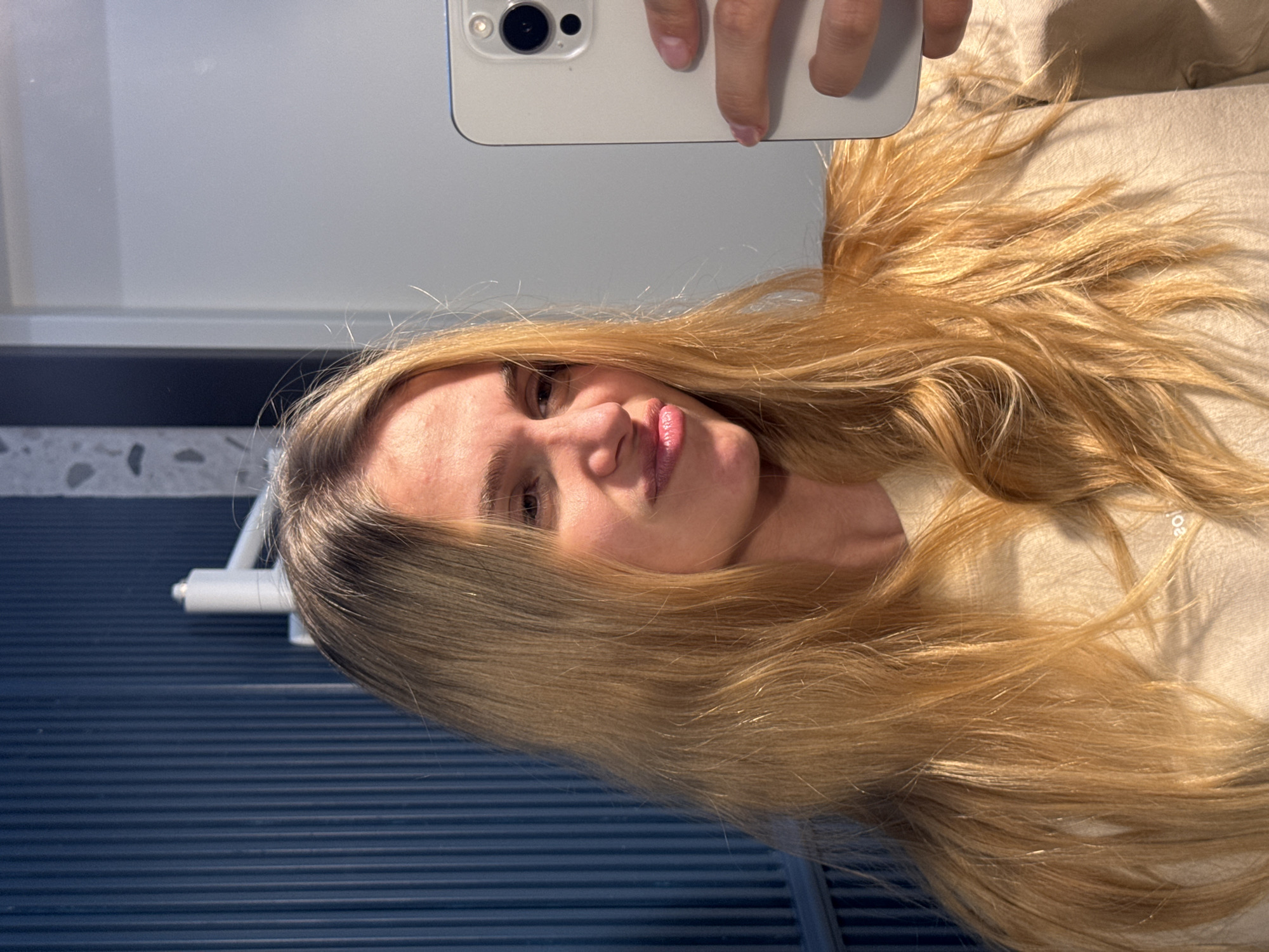 A selfie of a fair-skinned individual with long, blonde hair. The person is holding a white smartphone with a visible camera lens, presumably to capture the image. The background consists of blue, horizontally ridged surface and a wall with a patterned design. The person is wearing a light-colored top. The location is unknown.