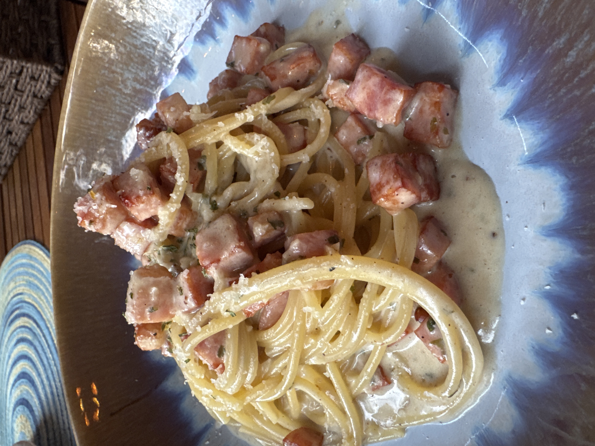 A close-up shot shows a plate of spaghetti carbonara. The spaghetti is coated in a creamy sauce, interspersed with small cubes of what appears to be cooked ham or bacon. The plate has a textured surface, with a light brown interior and a blue and white marbled exterior. The dish is placed on a table with a woven, natural-fiber mat to the left. The location is Baan Mae Nam, Thailand.